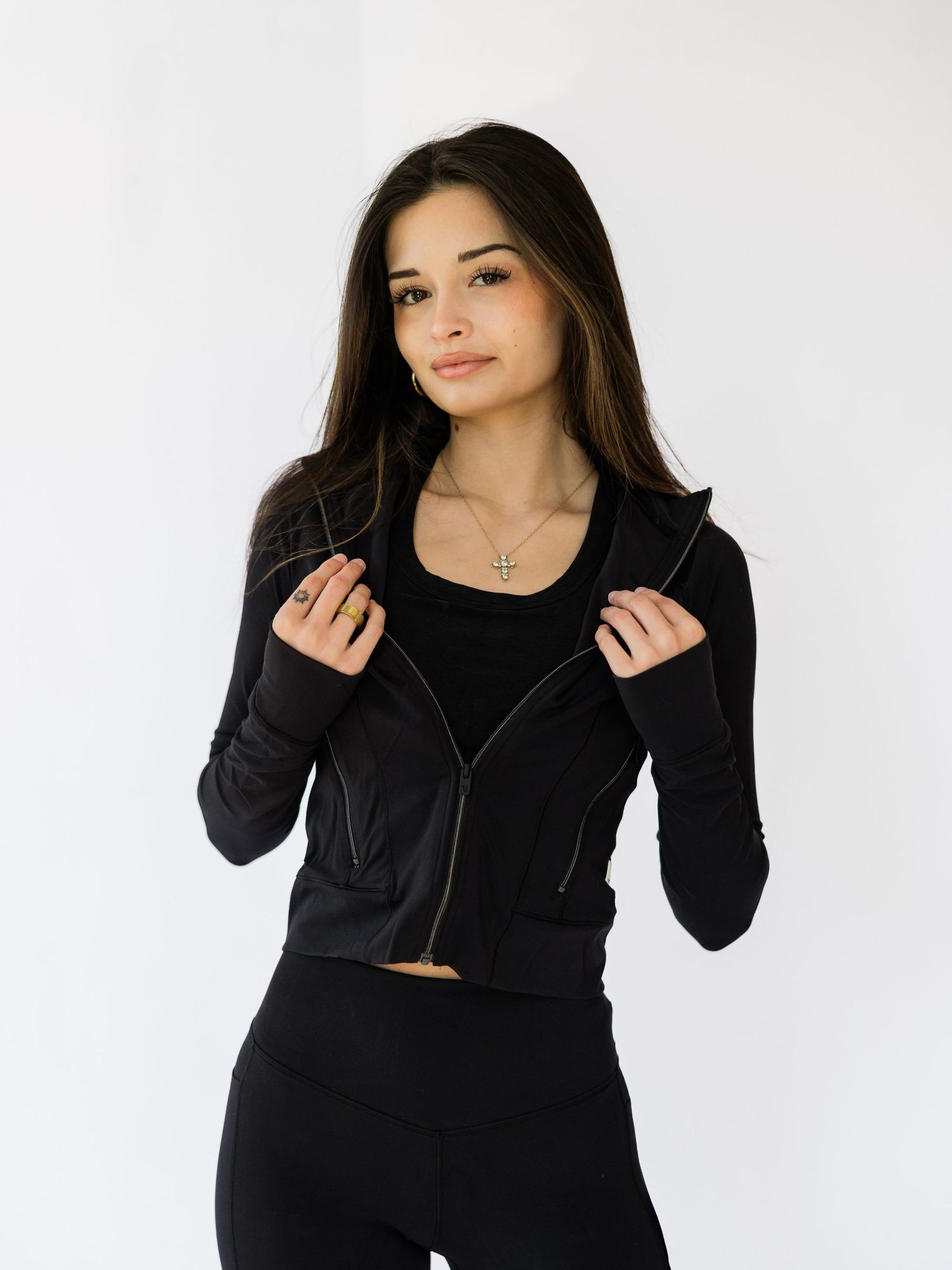 Woman wearing a black cropped moto jacket with zipper detail, black tank top, and leggings, styled with a cross necklace.