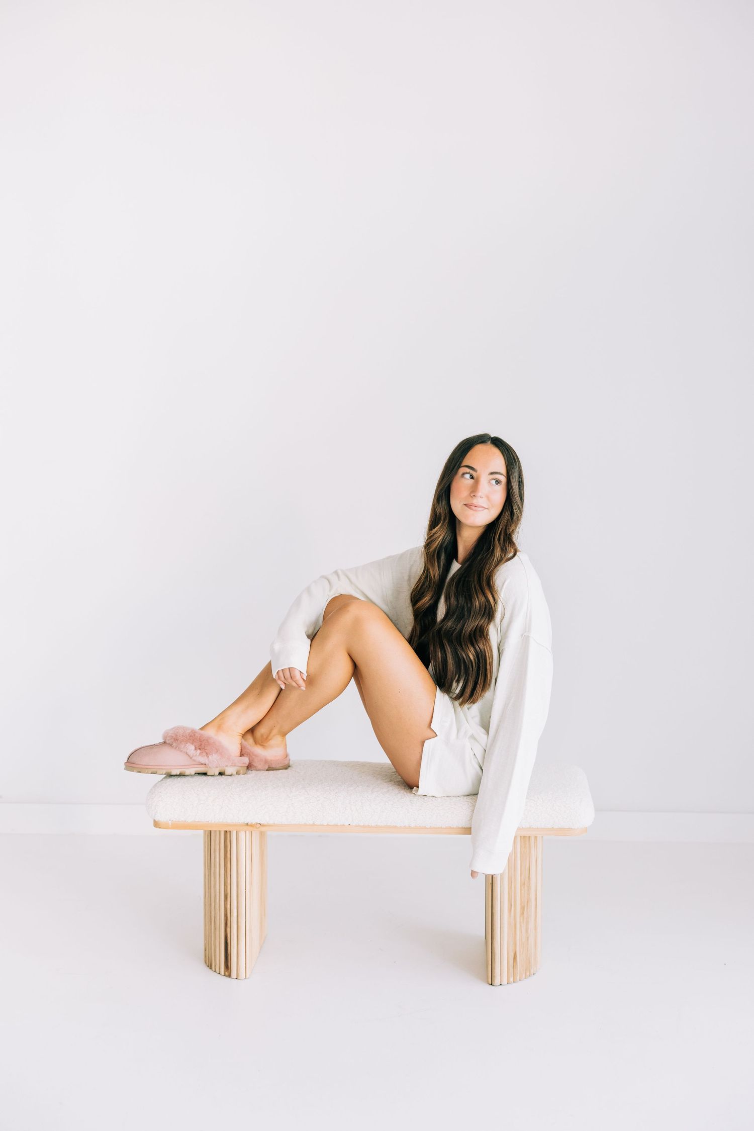 Woman in white oversized sweater and pink slippers sitting on a wooden bench against a white background.