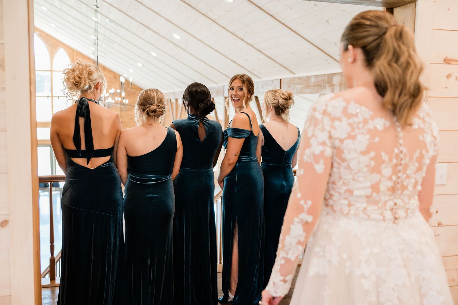A row of bridesmaids in black dresses stand together at a wedding venue with white walls and windows.