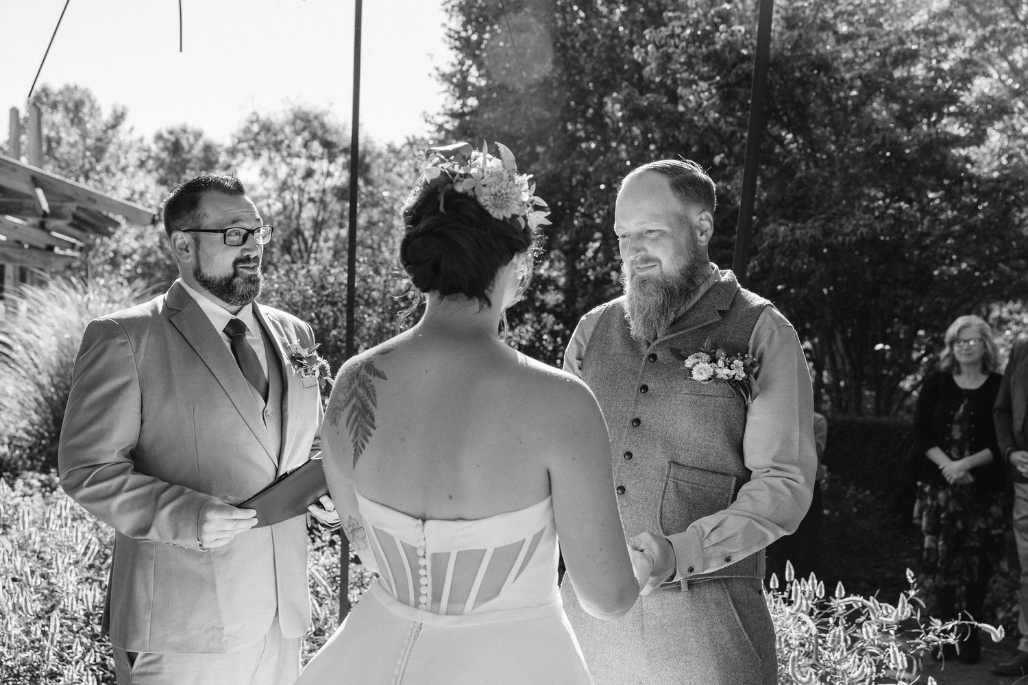 A black and white outdoor wedding ceremony shows a couple exchanging vows with an officiant in a garden setting.