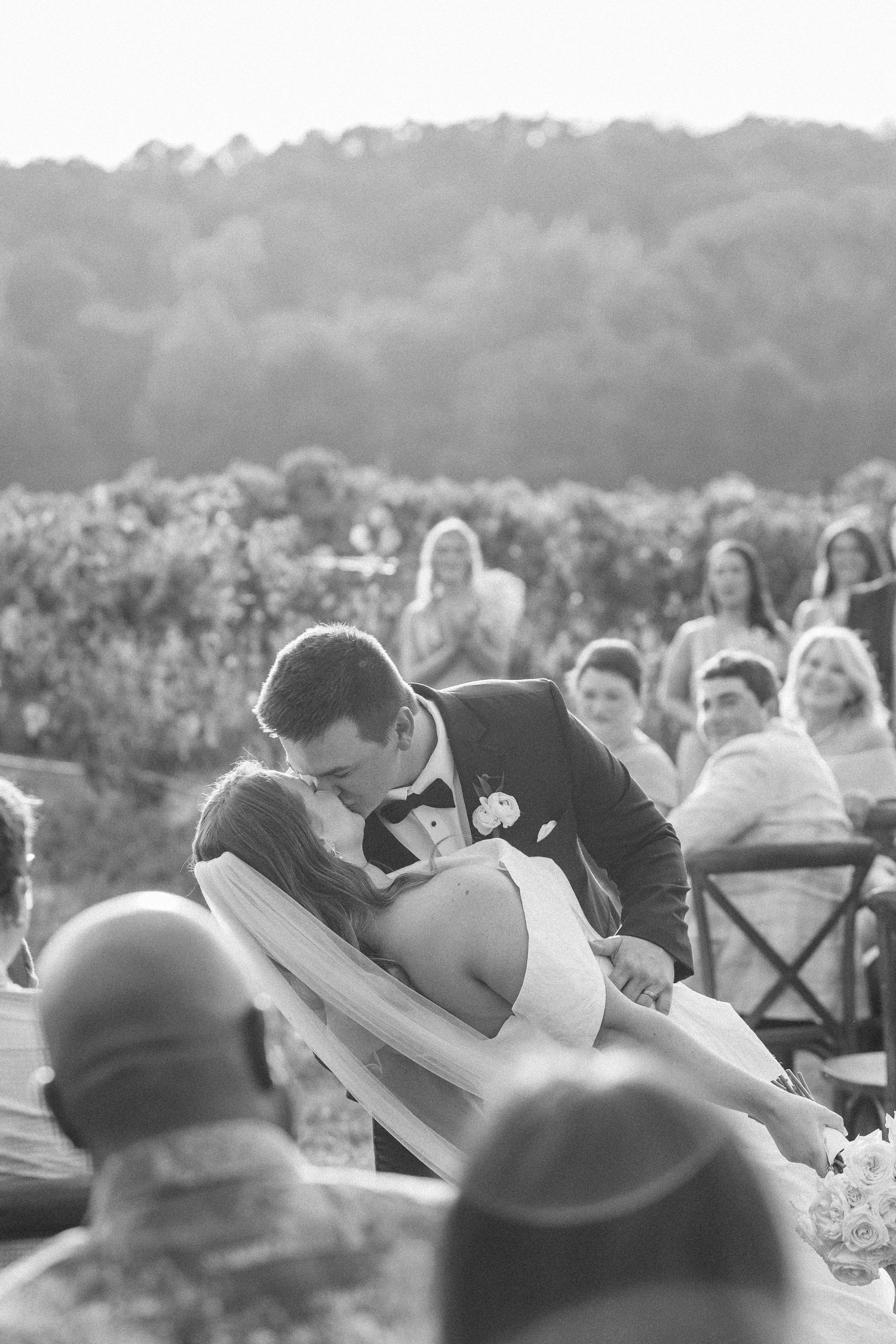 A romantic wedding kiss between newlyweds at an outdoor ceremony with guests looking on in the background.