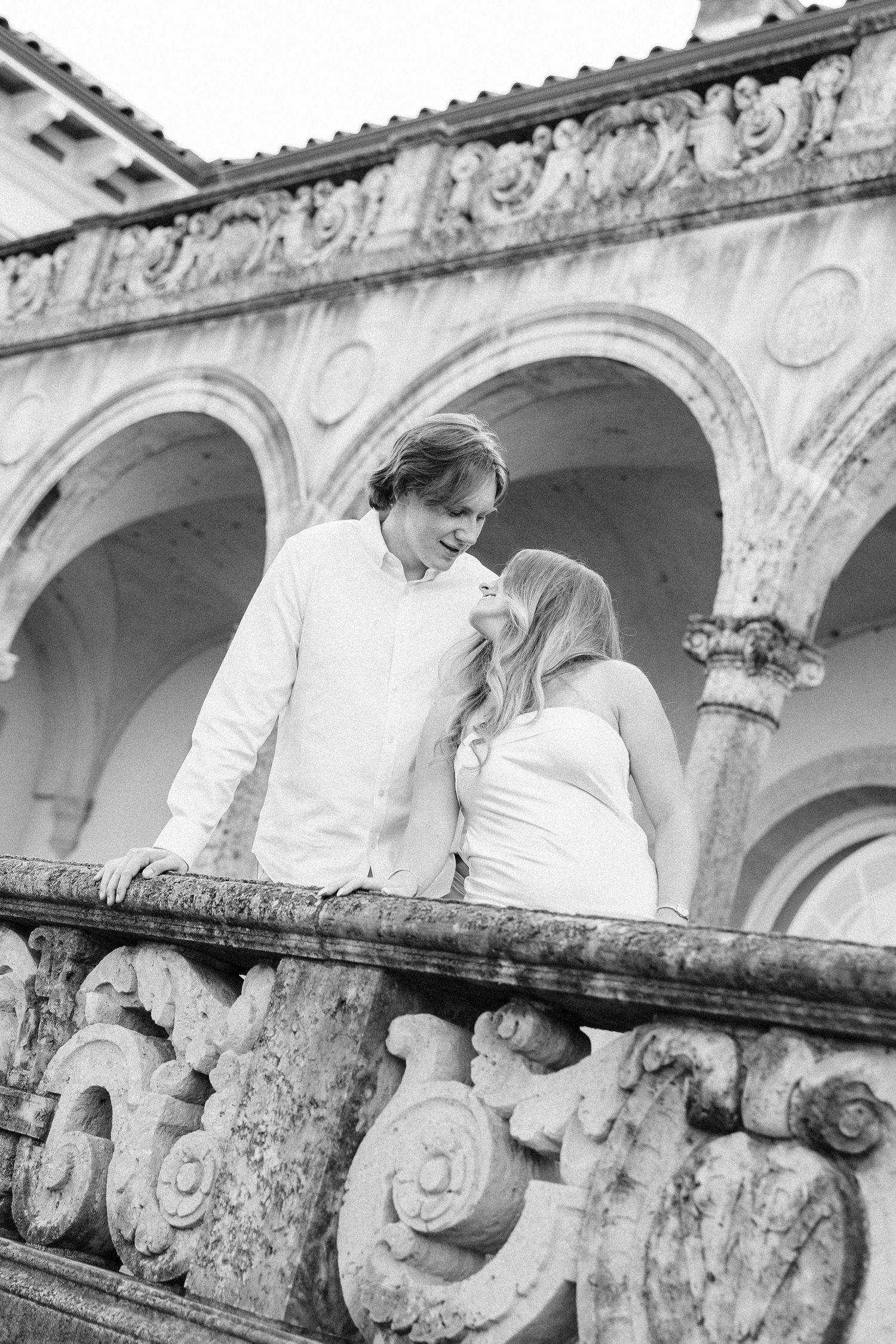 A person in white leans over an ornate stone balustrade with decorative carved scrollwork at a historic building.