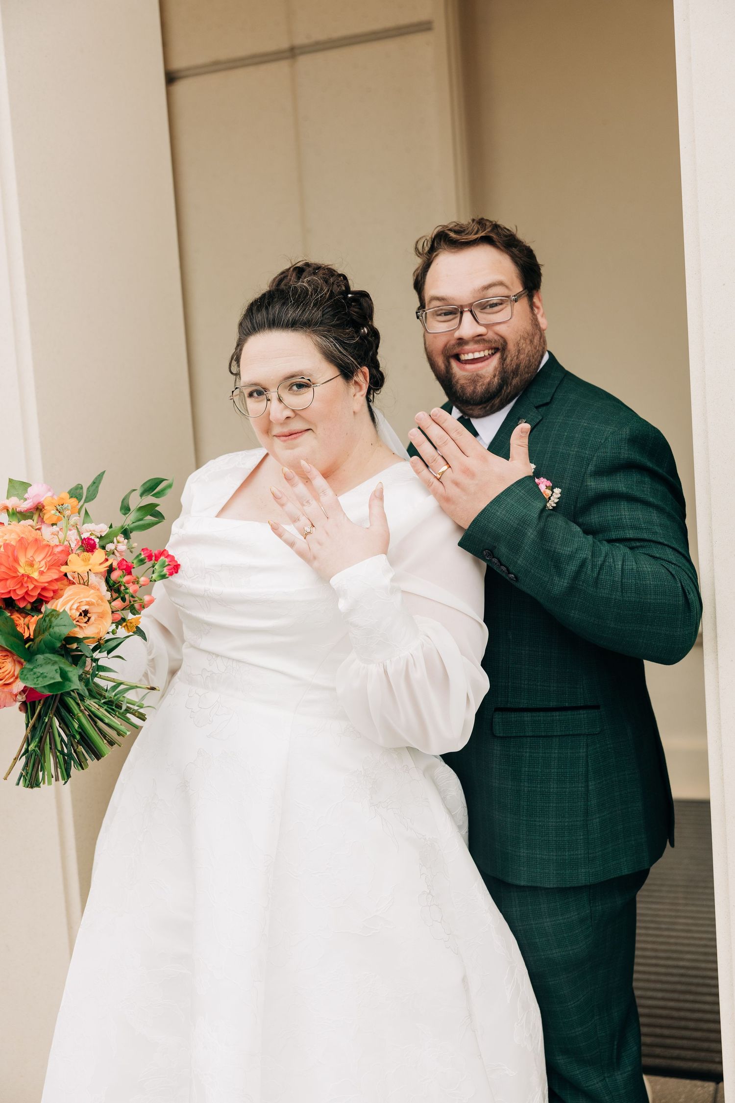 Newlyweds show off their rings while holding a vibrant orange and pink bouquet.