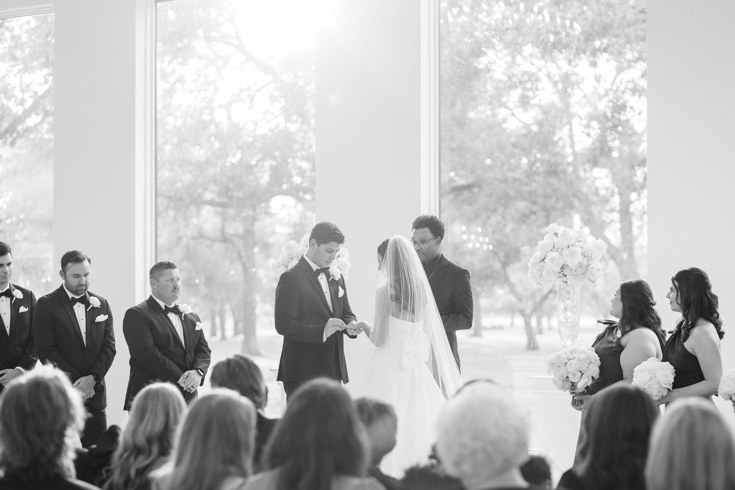 A black and white wedding ceremony scene with guests watching as a couple exchanges vows at an elegant indoor venue.