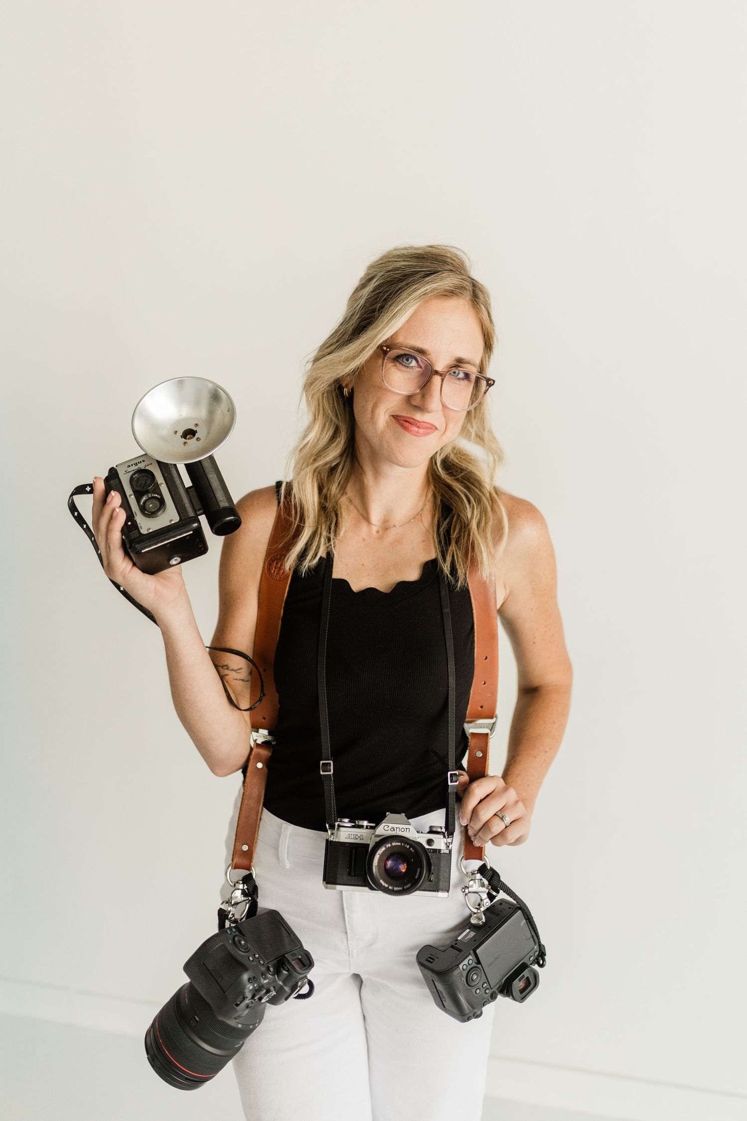 Woman photographer holding vintage and modern cameras, wearing glasses and black top with white pants, studio setting.