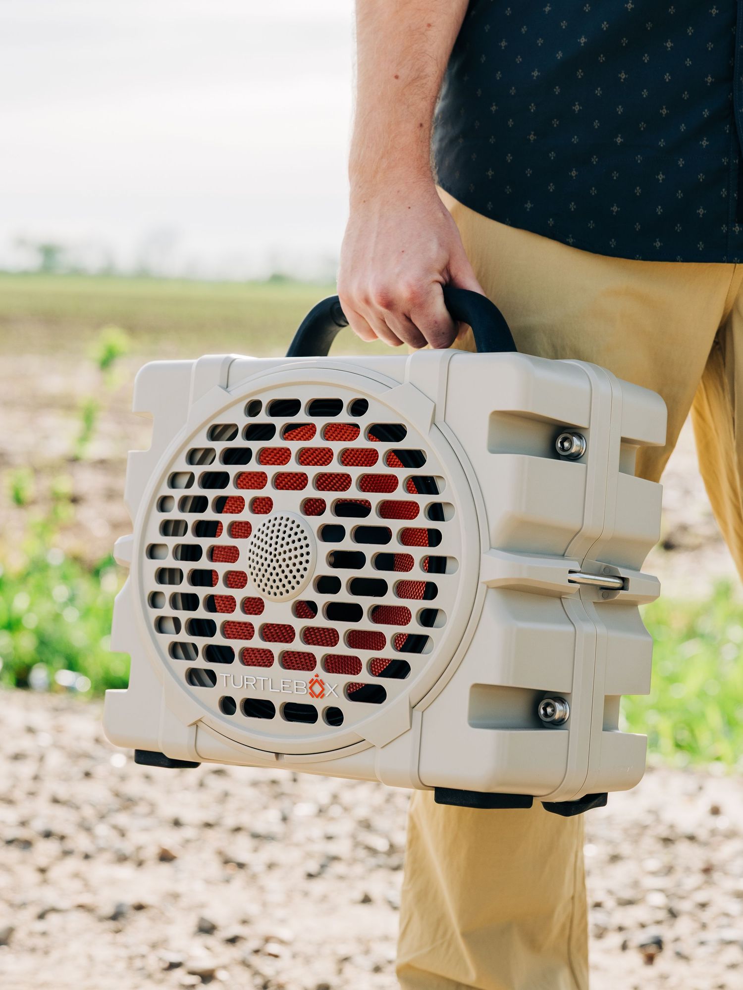 Person holding a rugged portable Bluetooth speaker with red grill accents outdoors in a field.