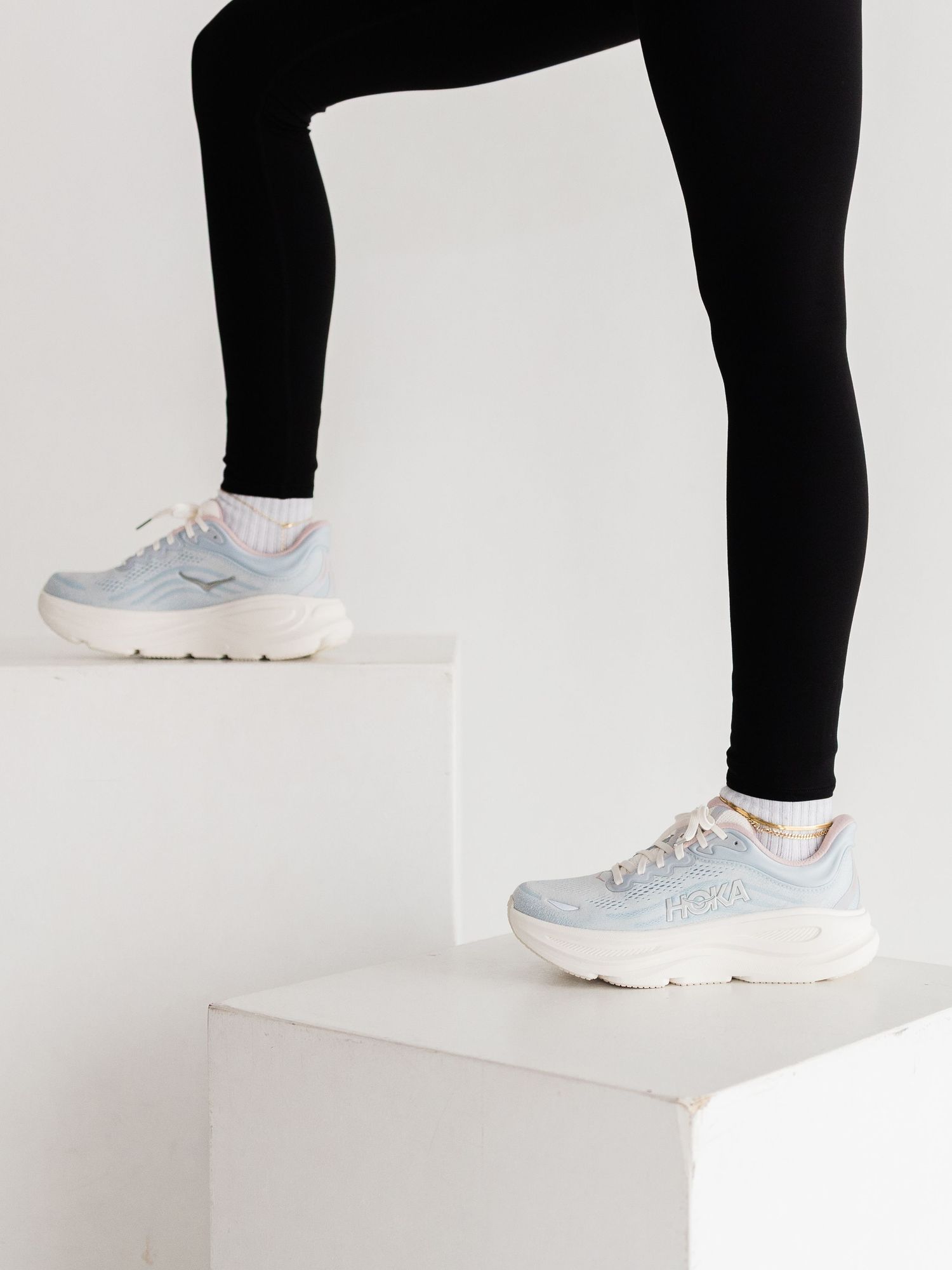 Person wearing black leggings and light blue chunky sneakers standing on white blocks against a white background.