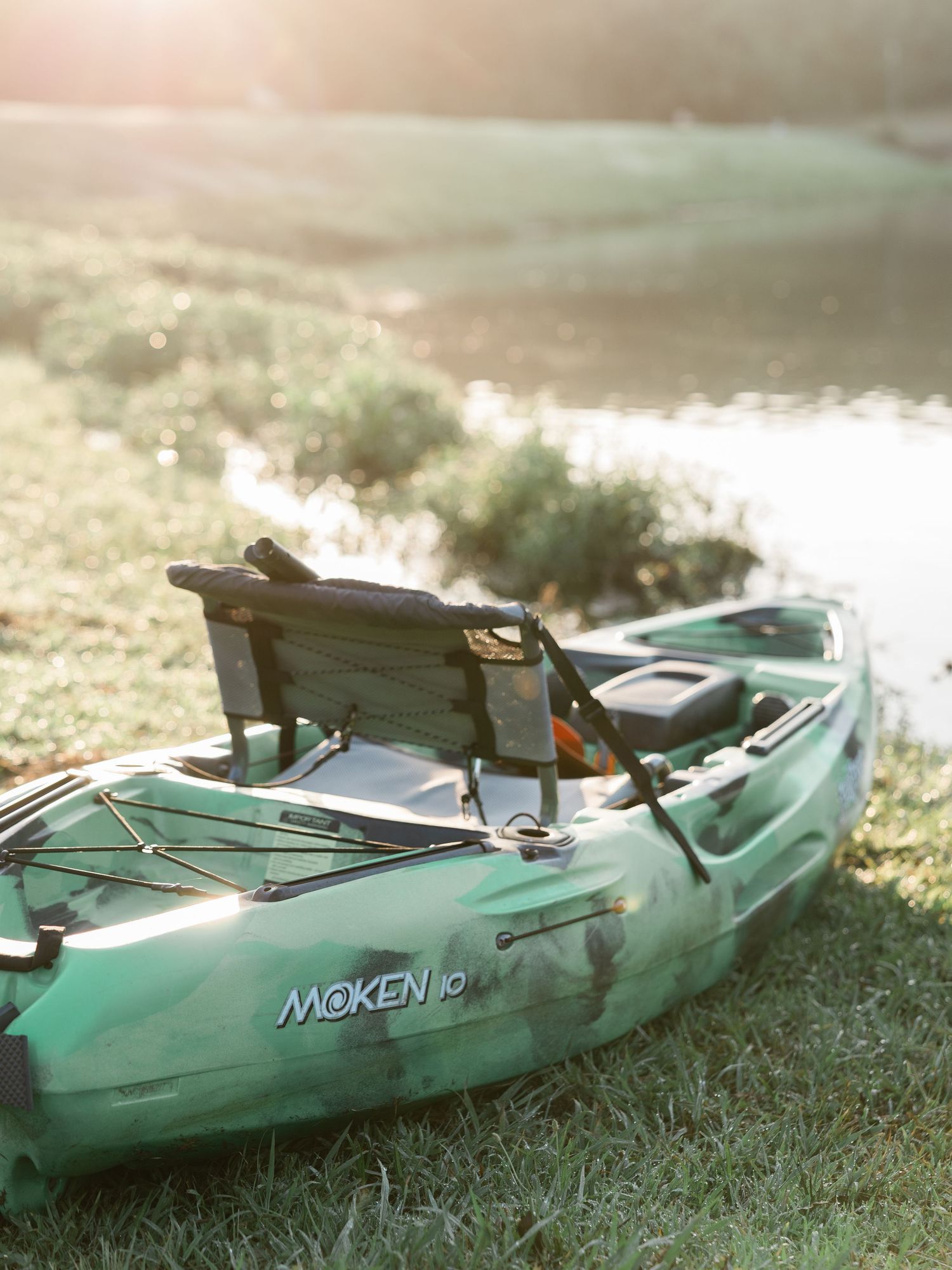 Green Moken 10 sit-on-top kayak resting on grassy riverbank at golden hour sunset.