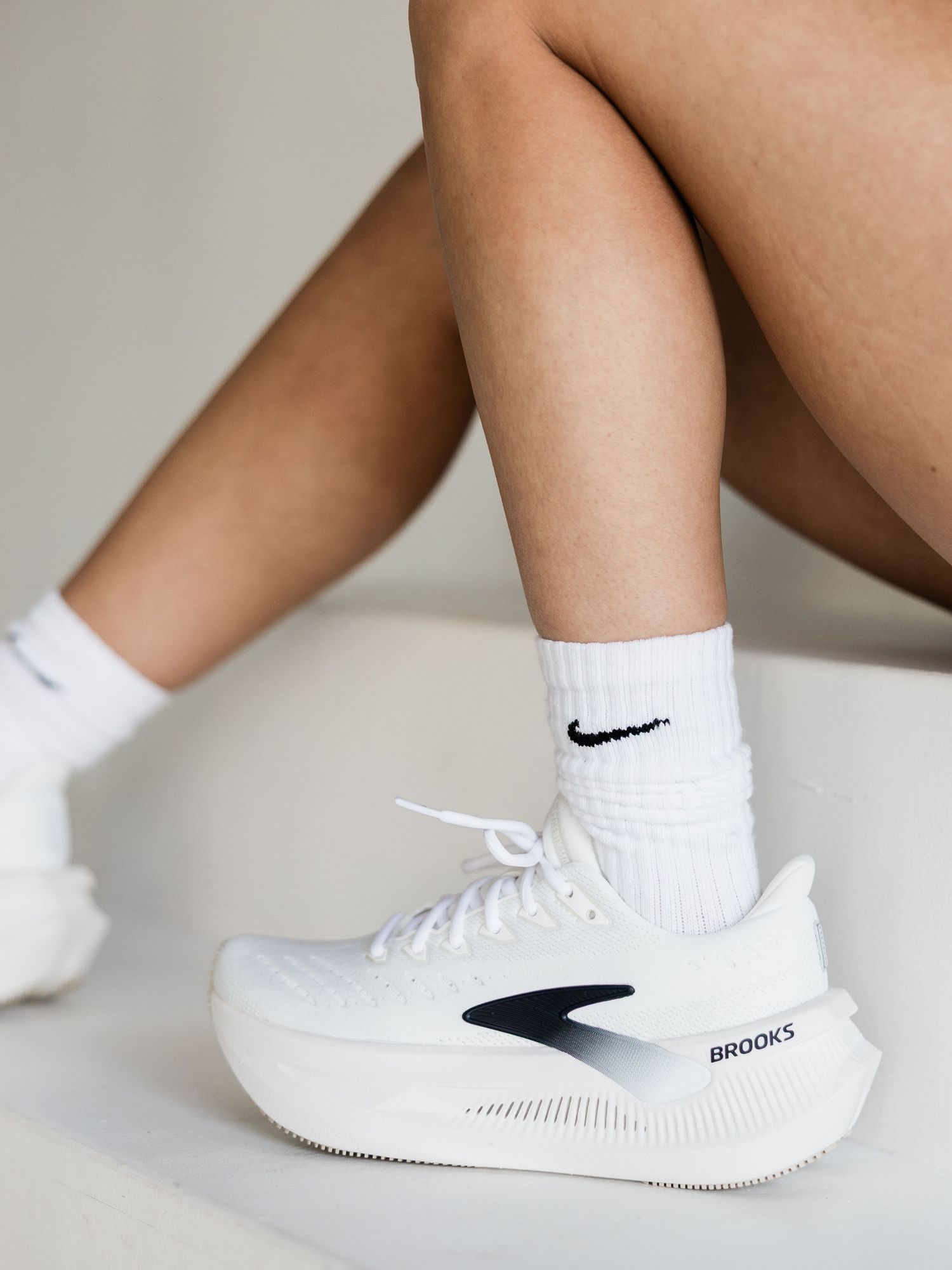 White Brooks running shoes with Nike socks on a seated person against a light background.