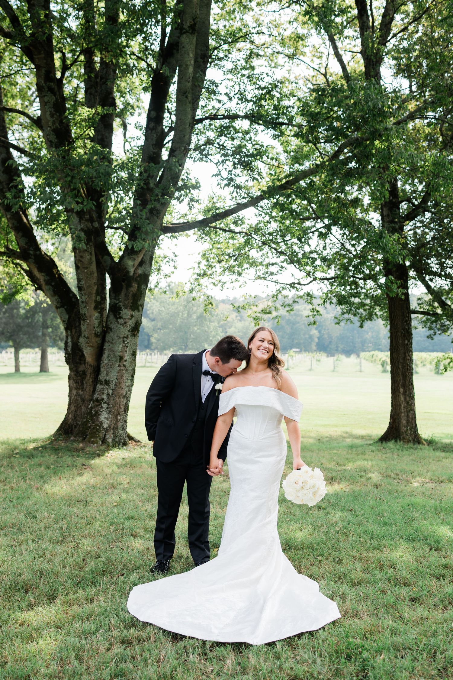 A couple shares a romantic moment under large oak trees in a grassy field during their wedding photo session.