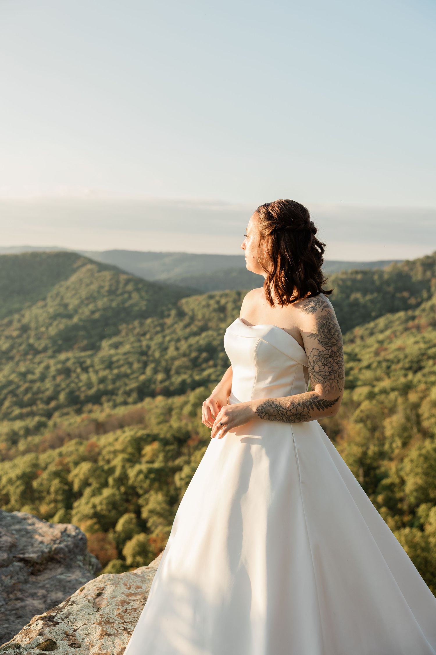 A bride in a flowing white ballgown stands on a mountain overlook during sunset with rolling hills in the background.
