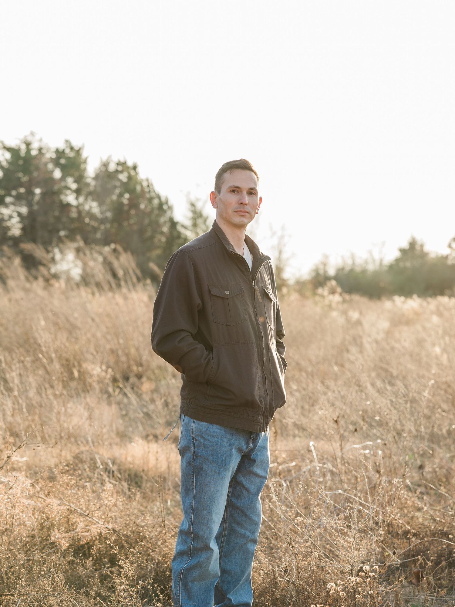 Young man in dark jacket and jeans standing in golden field with trees in background during sunset.