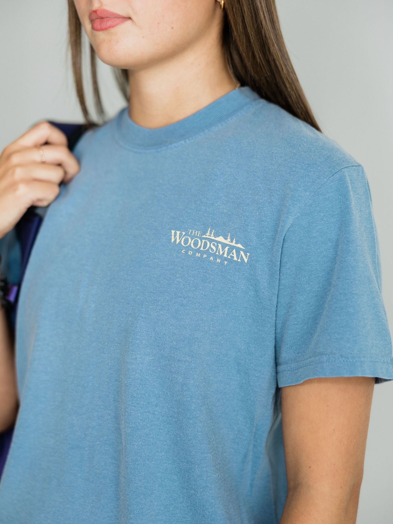 Woman wearing a blue The Woodsman Company logo t-shirt, close-up front view showing chest graphic detail.
