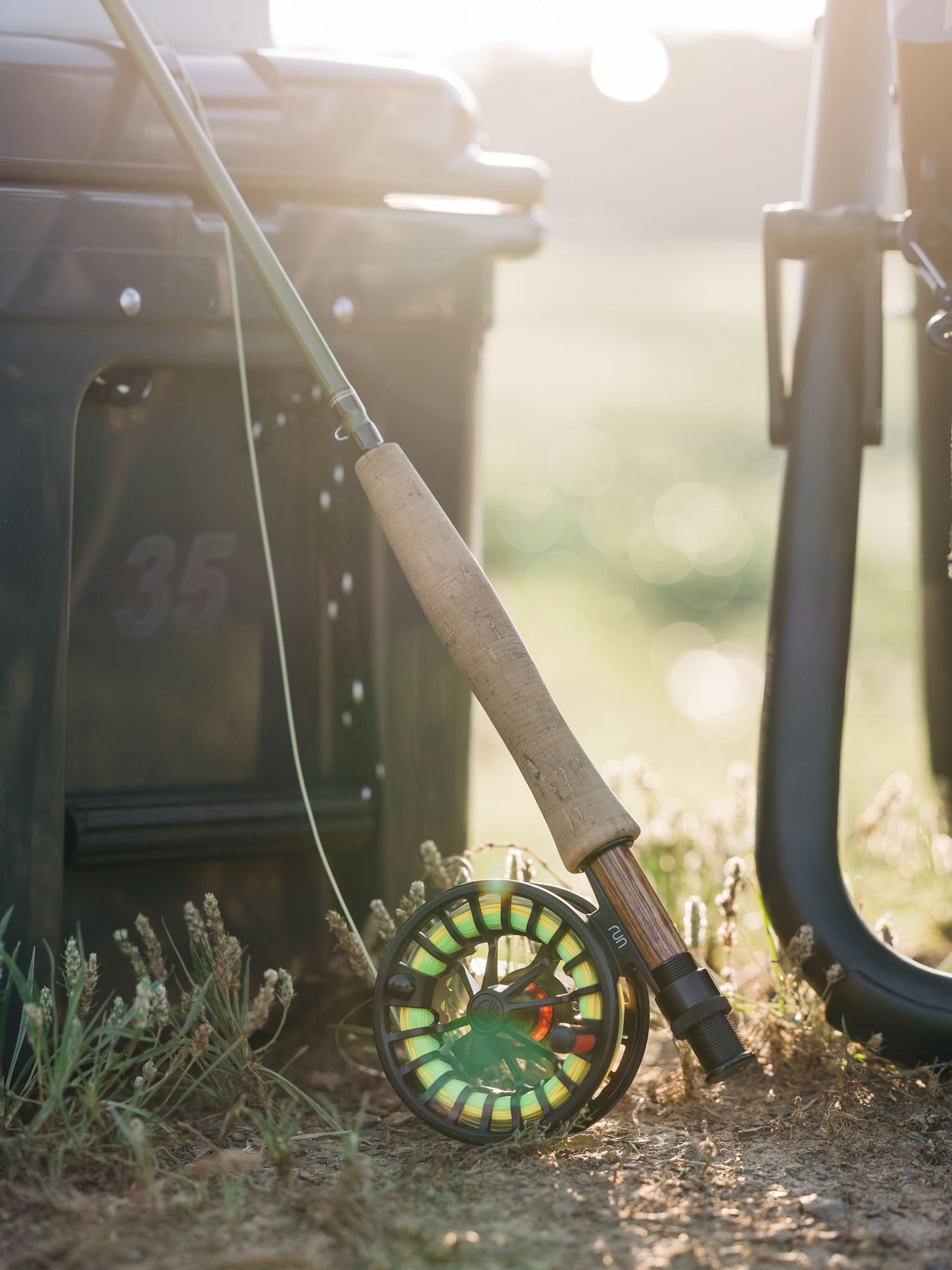 A colorful fly fishing reel and cork-handled rod leaning against dark green bins in warm golden sunlight.