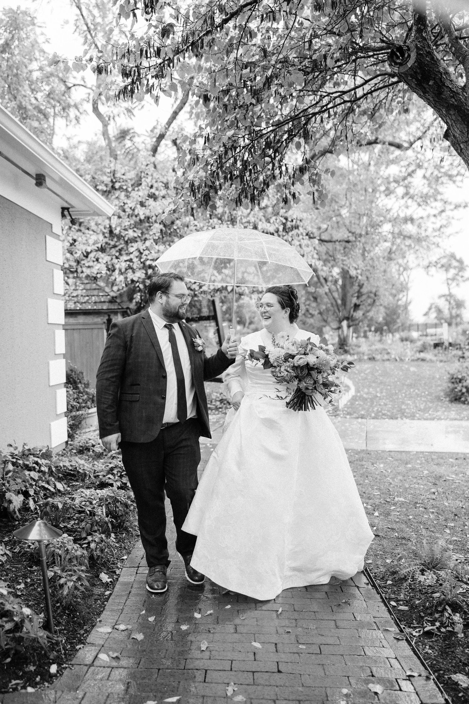 Couple walks along brick path with clear umbrella and colorful bouquet in black and white photo.
