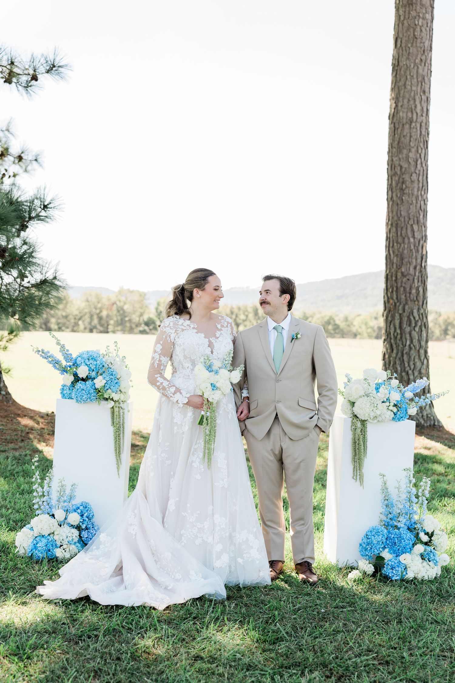 Wedding couple poses under pine trees with blue and white floral decorations.