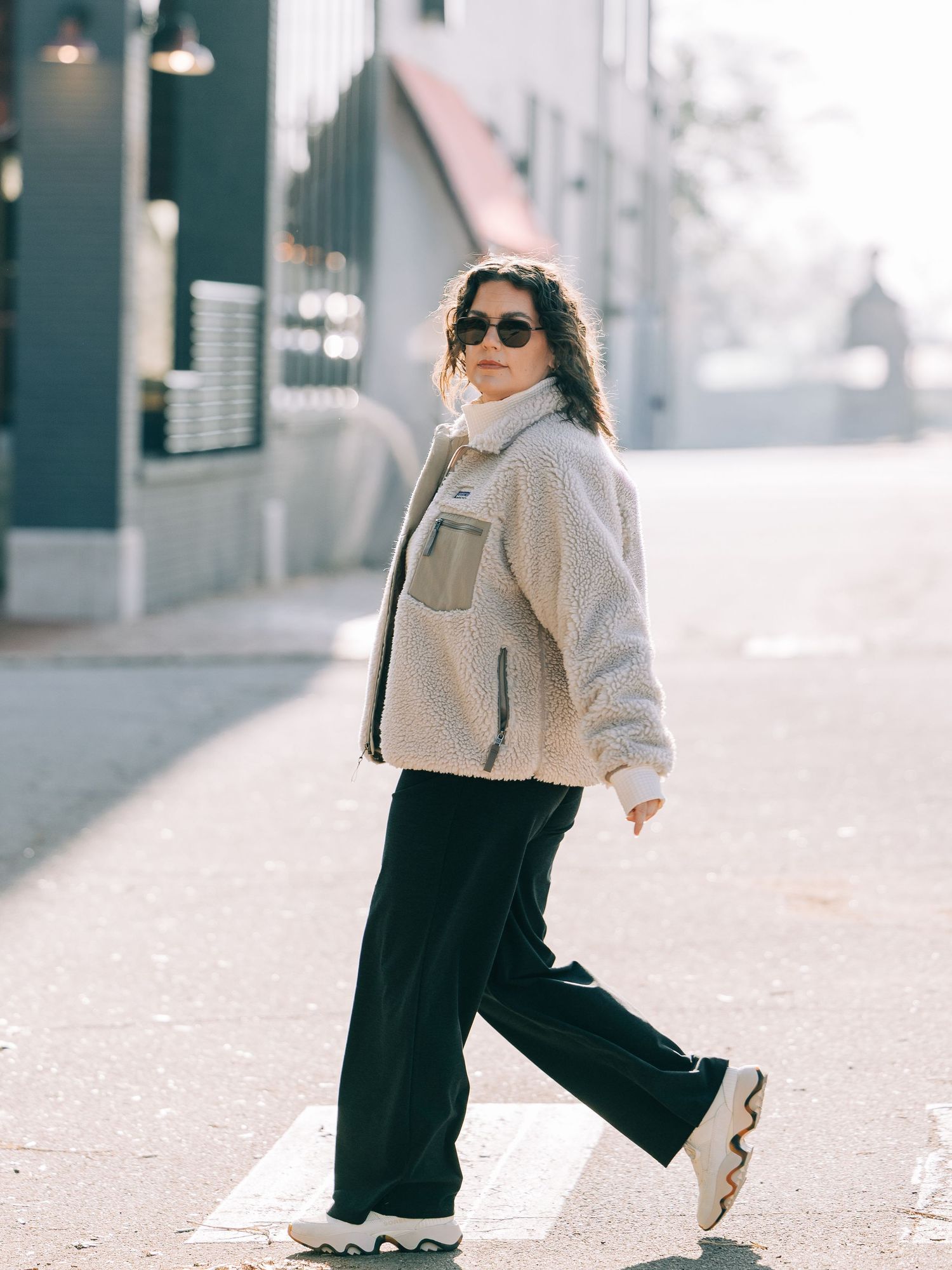 Woman in sherpa jacket, black wide-leg pants, and chunky sneakers walking on a sunny urban street.