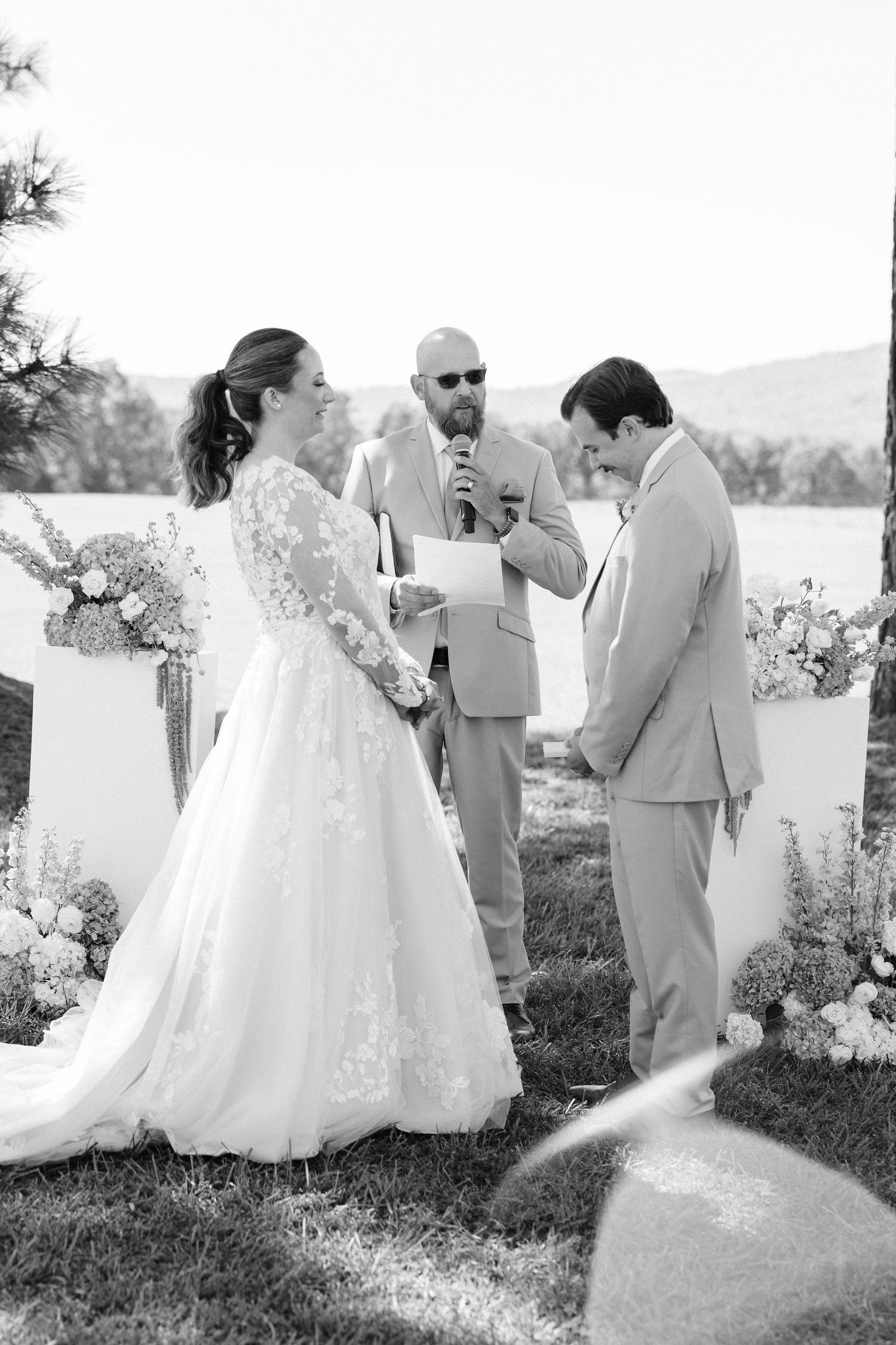 Wedding party gathers outdoors for ceremony photos in light colored attire.