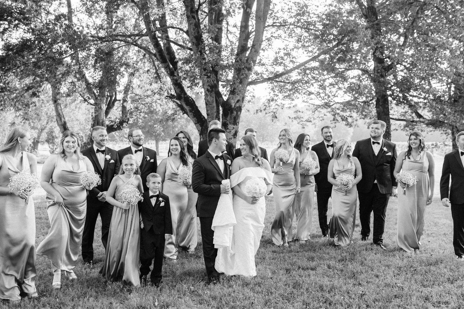 A wedding party stands in a line outdoors under trees wearing formal attire in black and white photography.