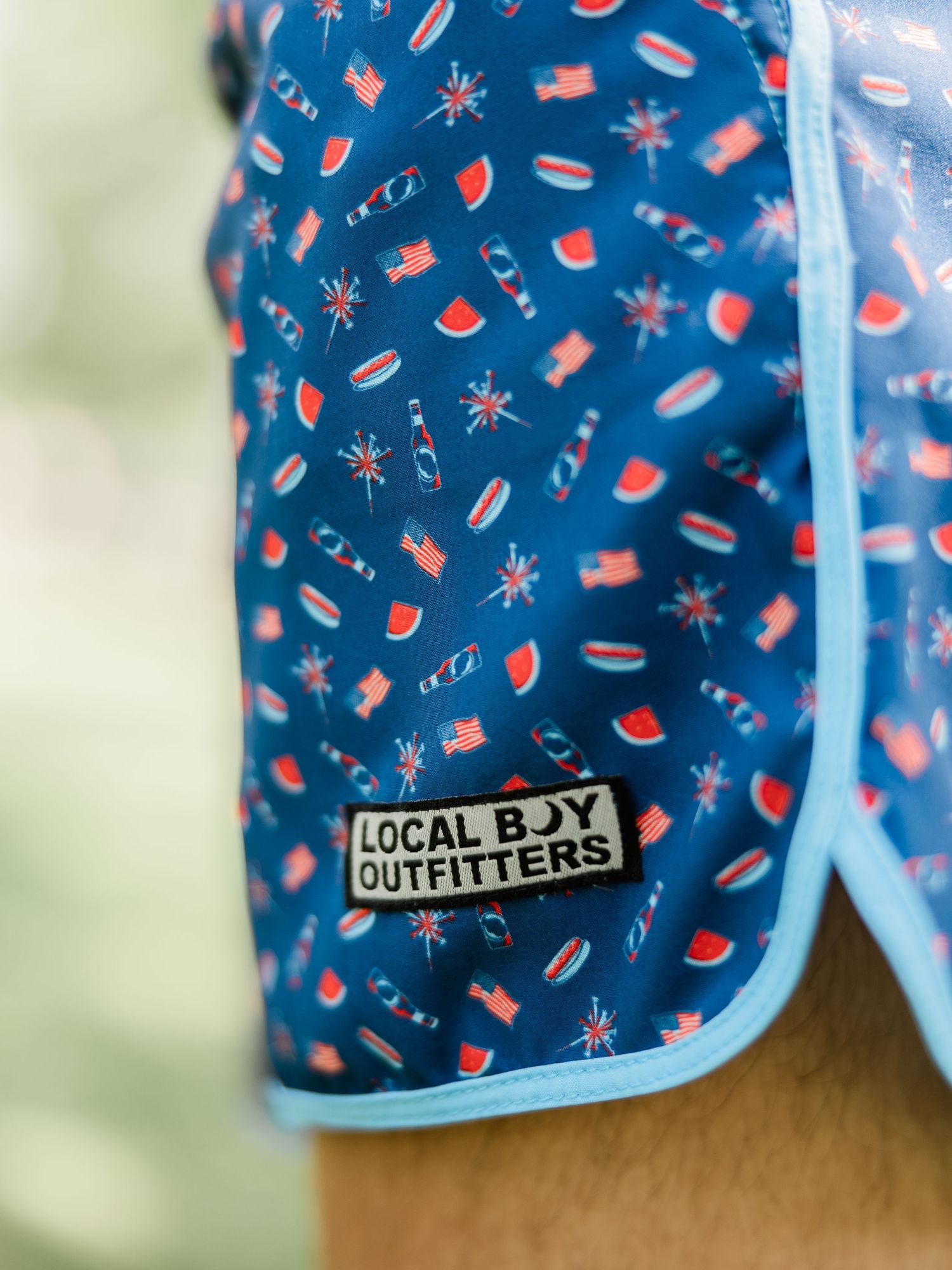 Close-up of Local Boy Outfitters blue patriotic swim trunks with American flags, watermelon, and fireworks print.