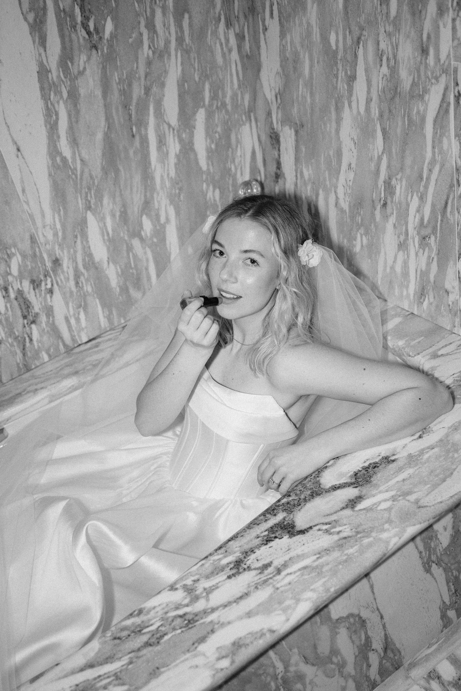 Black and white photo of someone reclining in a white corset dress against textured wall background.