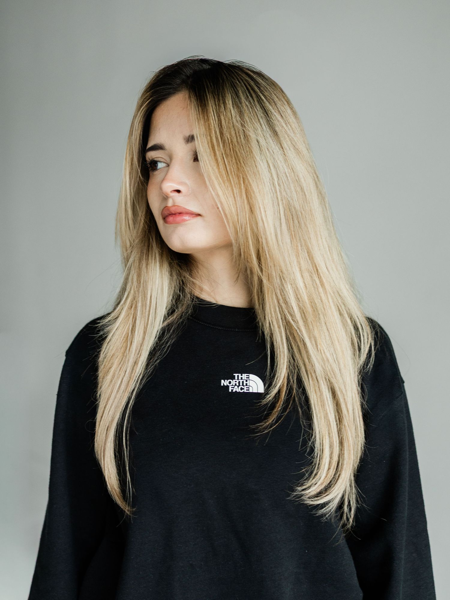 Young woman with long blonde hair wearing a black The North Face sweatshirt against a gray background.