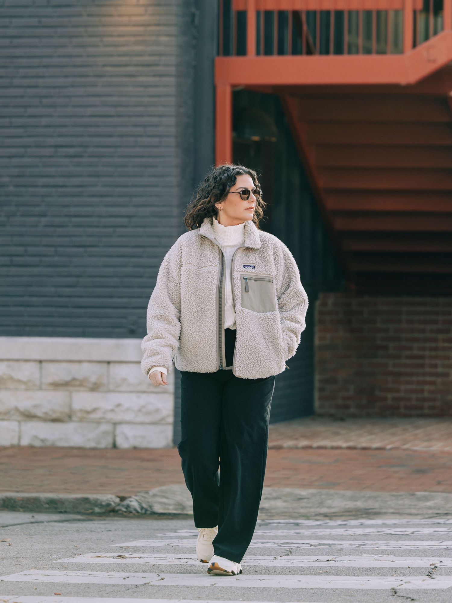 Woman in cream sherpa jacket and black pants crossing a street in an urban setting.