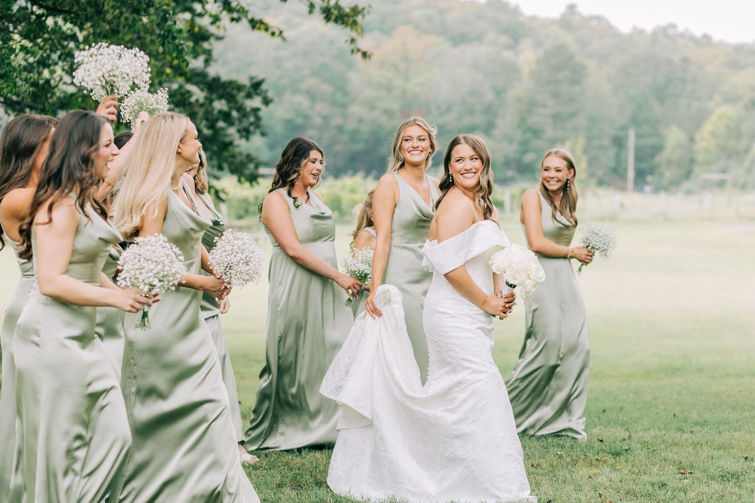 A bridal party in sage green bridesmaid dresses and white wedding gown pose playfully in a misty outdoor field.