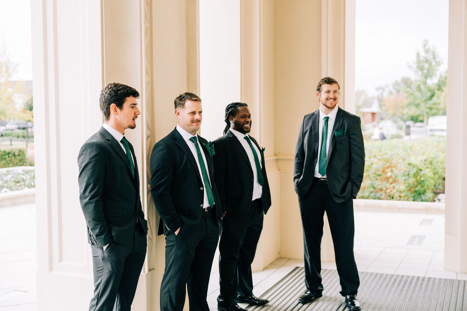 Groomsmen in black suits stand in line on a covered porch, creating a classic wedding party formation.