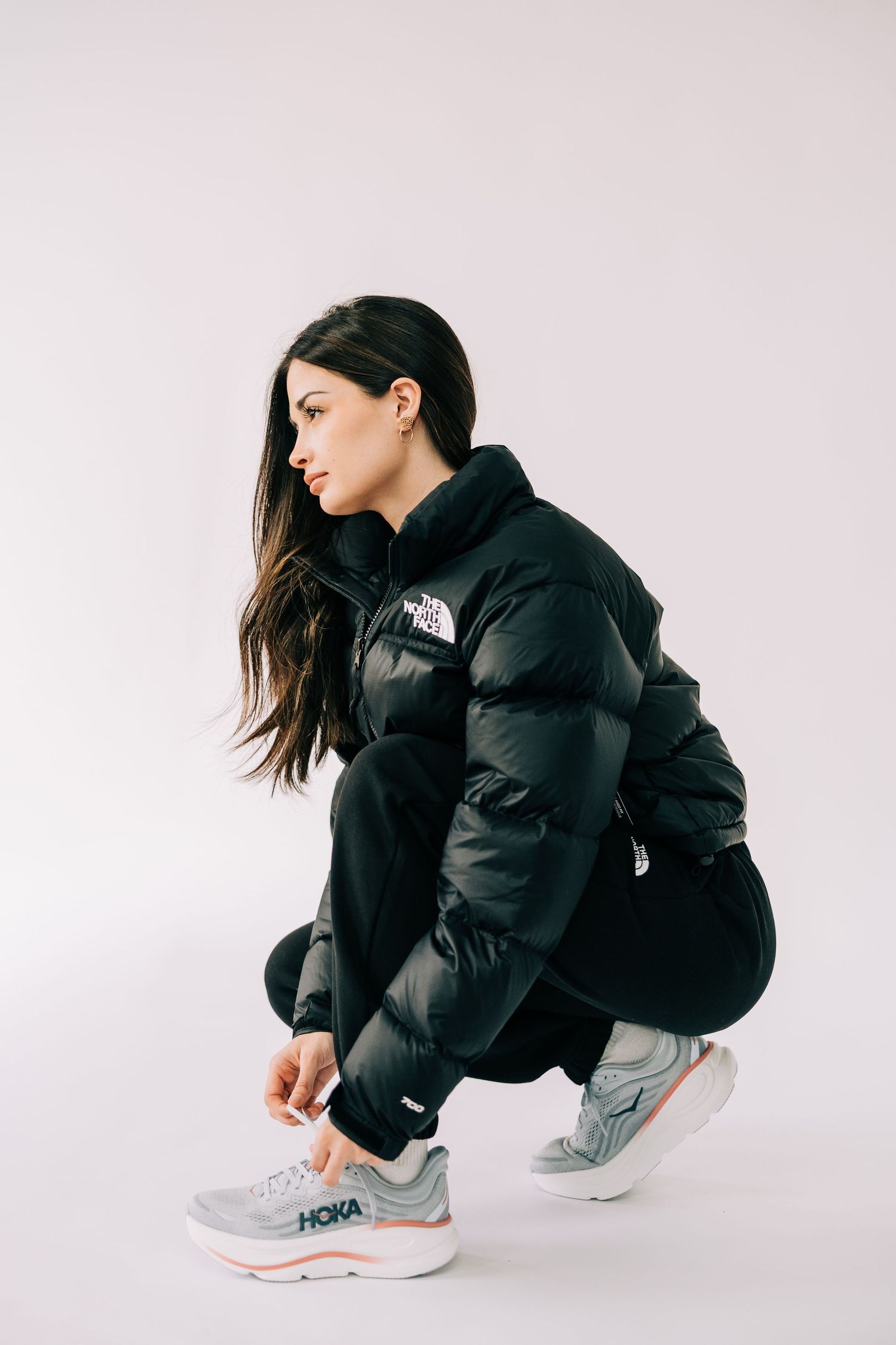 Athletic model wearing black North Face puffer jacket and gray sneakers crouches down in studio setting.
