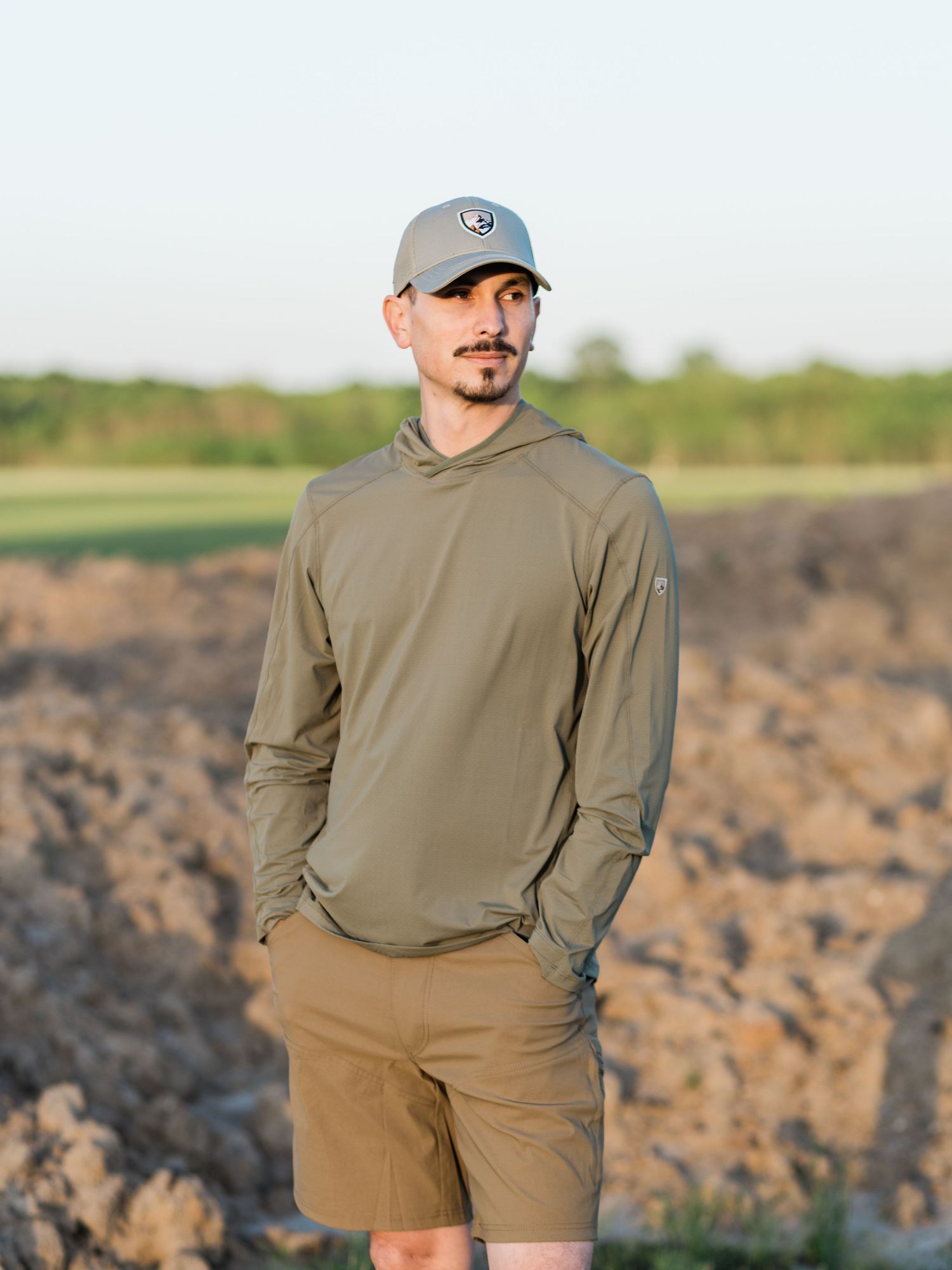 Man wearing olive green hoodie, khaki shorts, and gray cap standing outdoors in a field at dusk.
