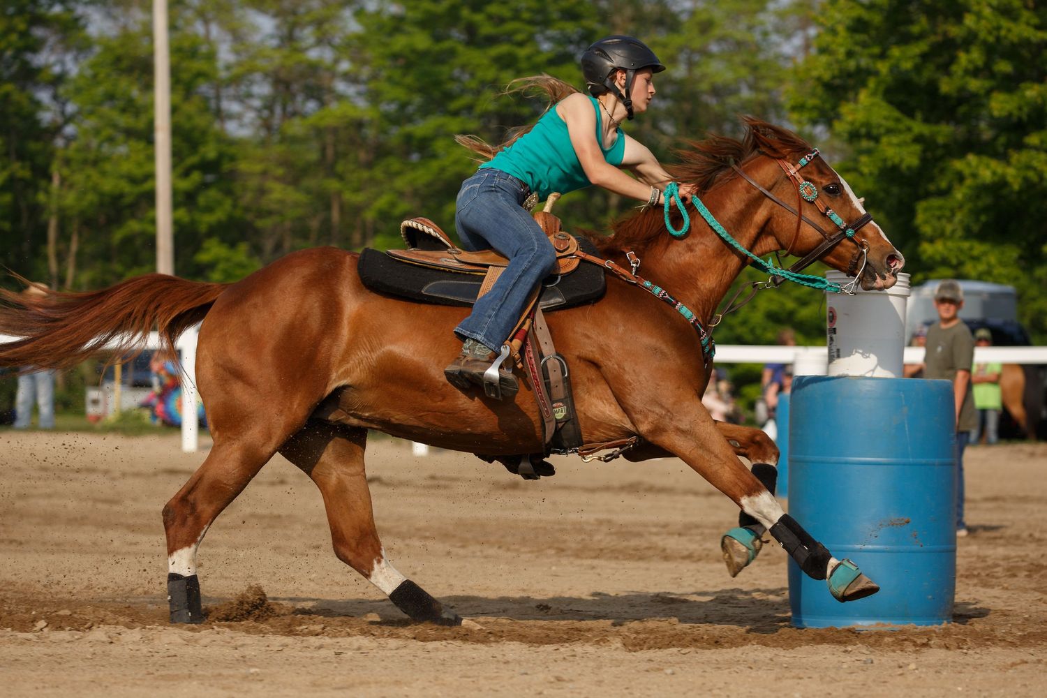 A rider on a chestnut horse moves at high speed while competing in a barrel racing event at an outdoor arena.