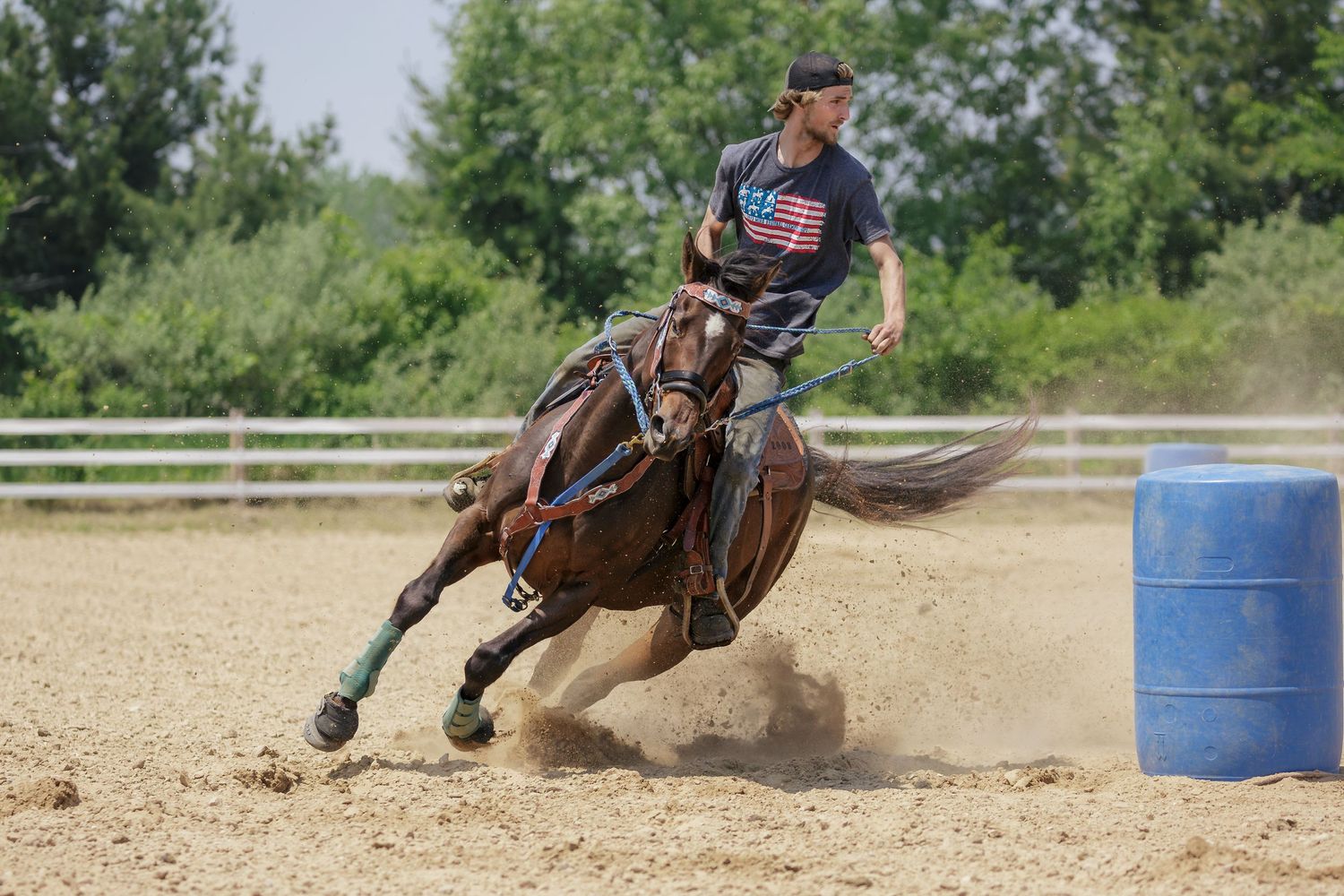 A rider on horseback races around a blue barrel in a dusty arena during barrel racing competition.