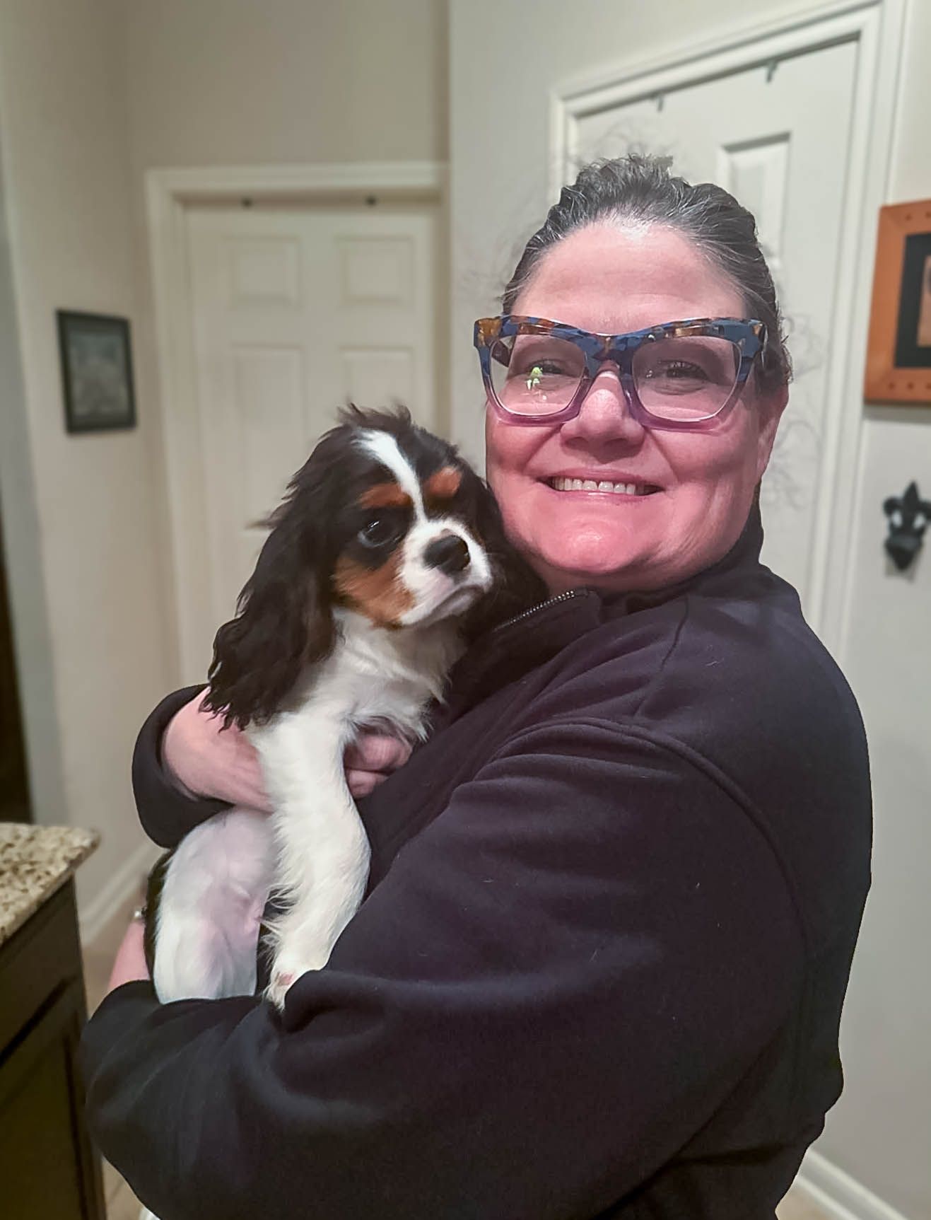 Smiling woman with colorful glasses holding a Cavalier King Charles Spaniel puppy indoors.