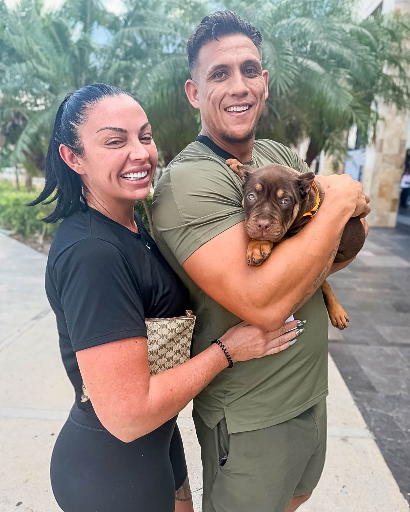 Smiling couple holding a small brown puppy outdoors near tropical palm trees.