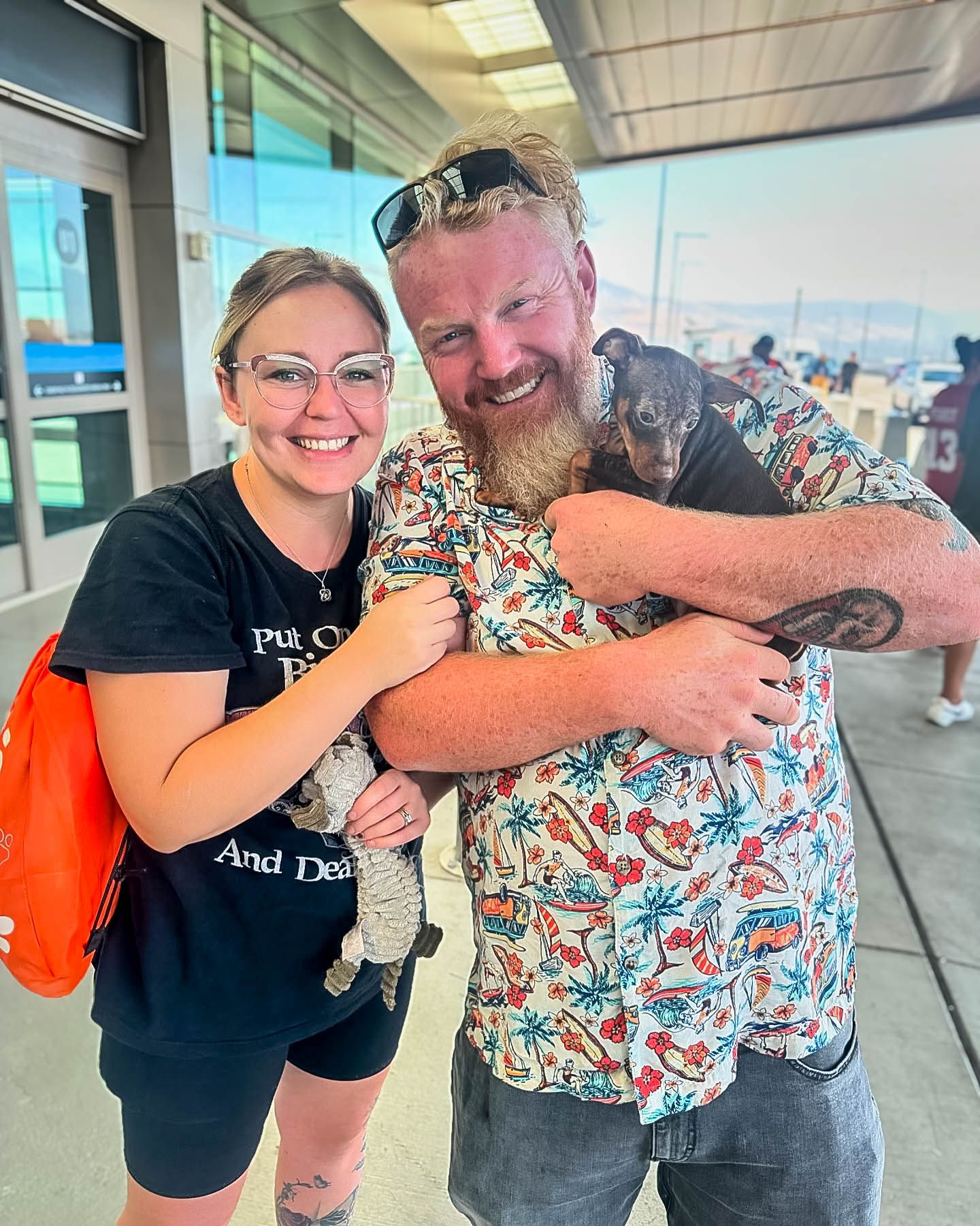 Smiling couple outside an airport embracing a small dark puppy together.