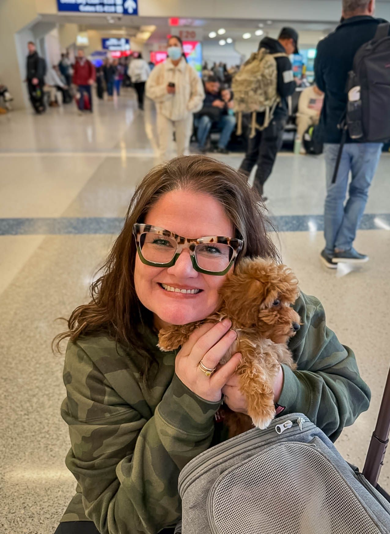 Woman smiling with small fluffy brown puppy in busy airport terminal, wearing green hoodie and leopard print glasses.