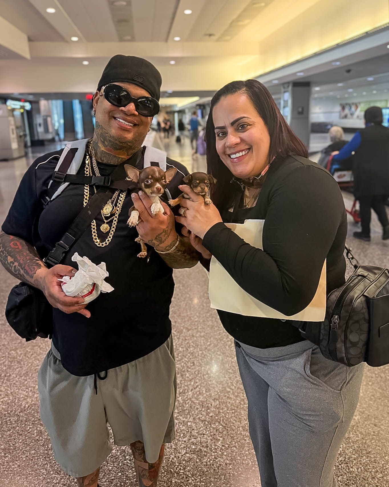 Smiling couple at airport holding two tiny Chihuahua puppies, man wearing gold chains and cap.