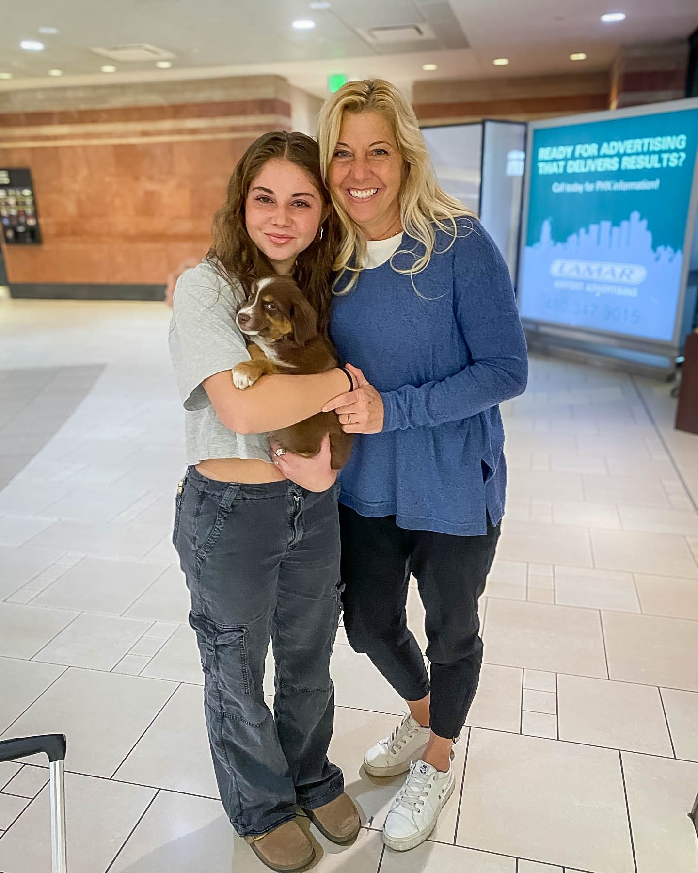 Two women smiling and posing together in an airport lobby, one holding a small brown dog.
