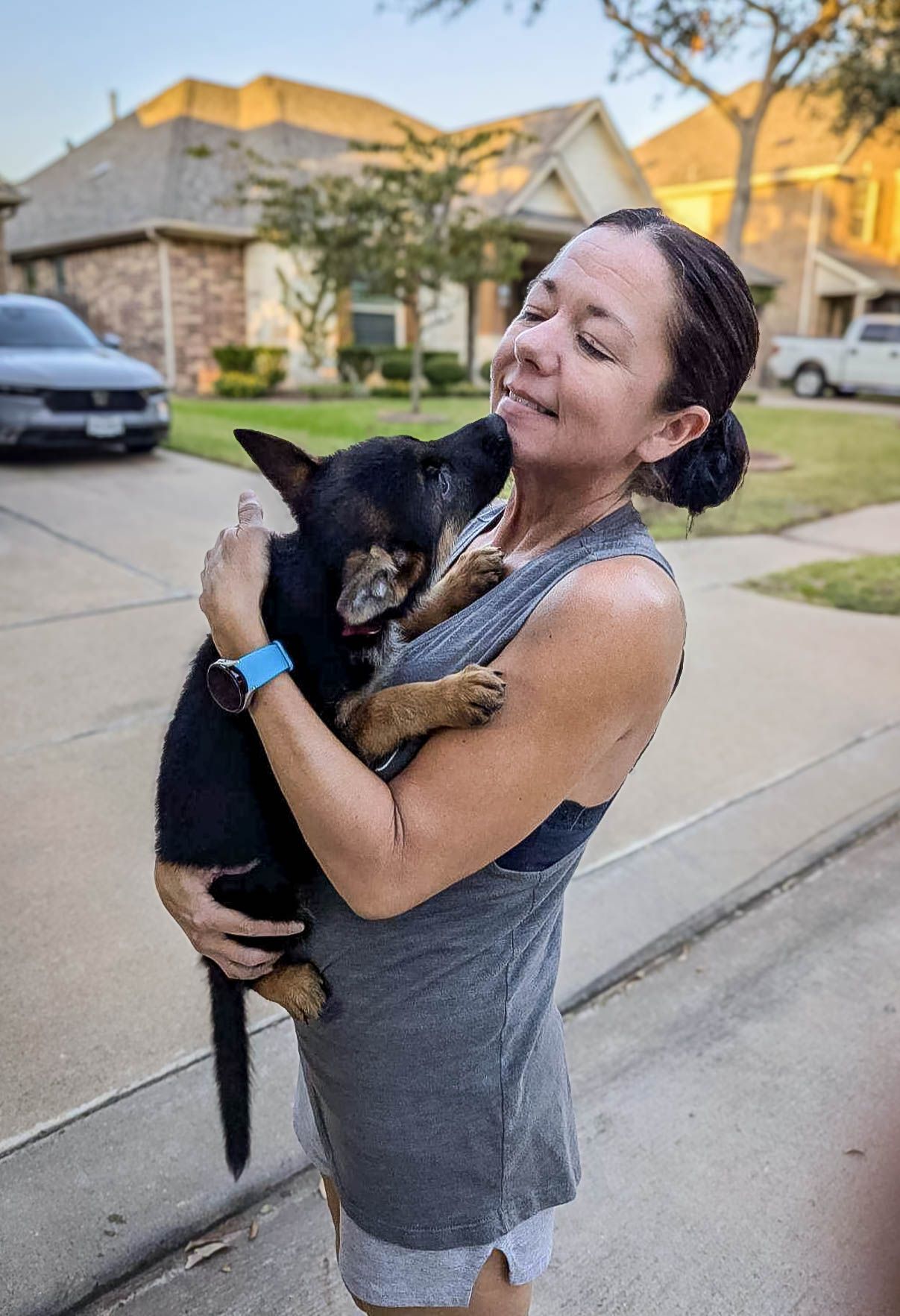 Woman joyfully cuddles a black and tan puppy on a suburban driveway at dusk after pet transport