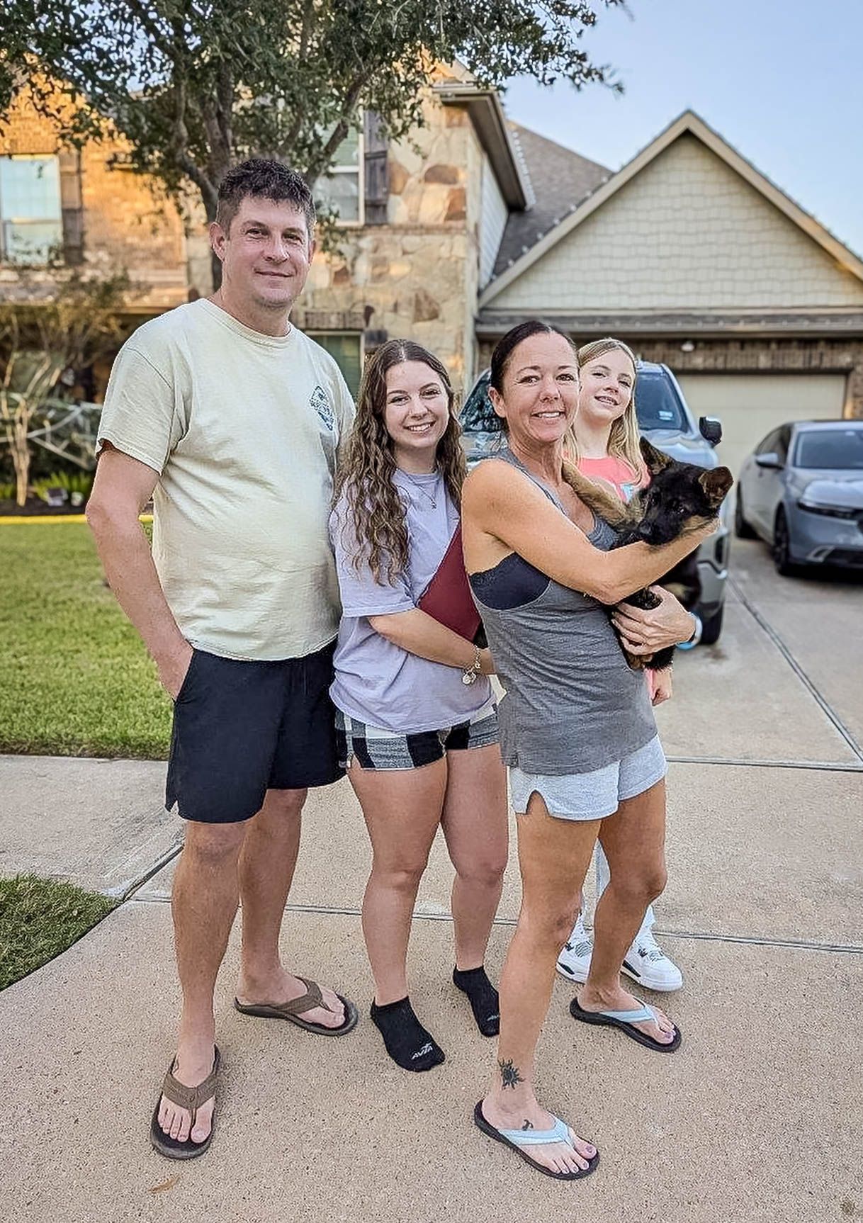 Family of four smiles in driveway holding a small black puppy outside suburban home, after pet transport