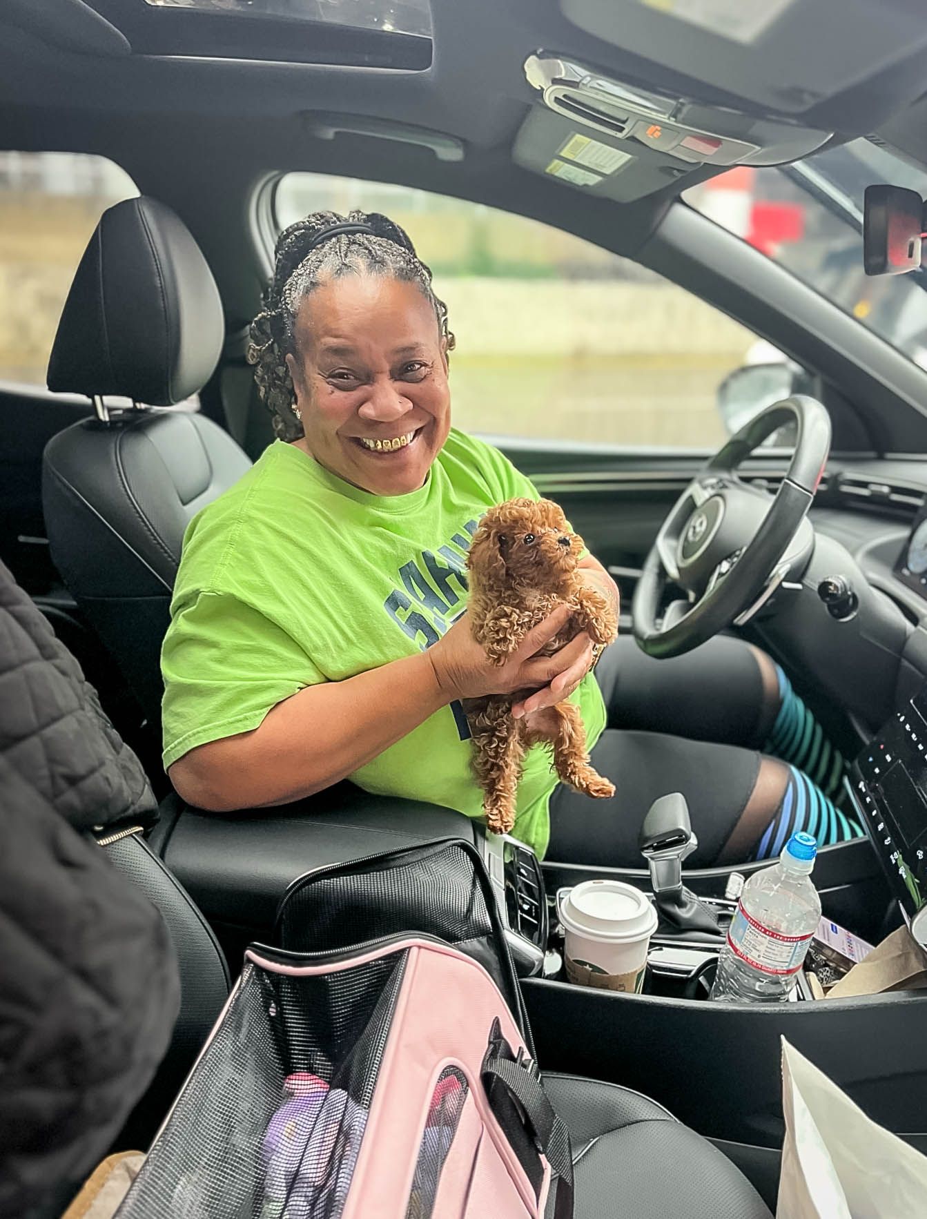 Happy woman in green shirt holds a red Toy Poodle puppy while sitting in a car after pet transport