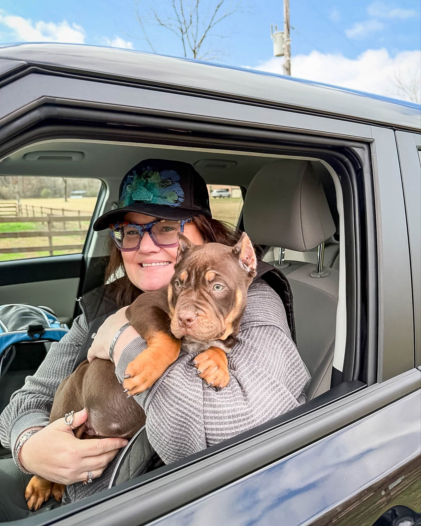 Woman smiling in car window holding a brown pitbull puppy on a sunny day during pet ground transport.