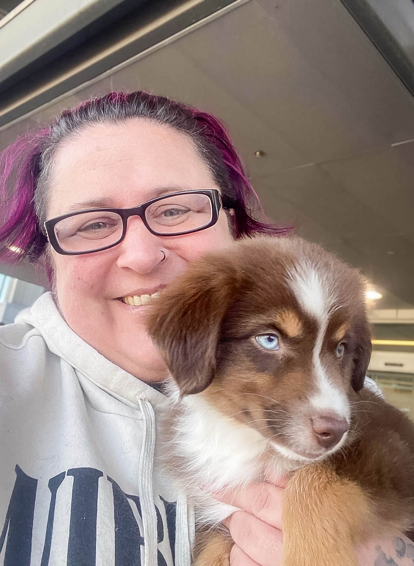 Woman with purple hair and glasses holding a brown and white Australian Shepherd puppy.