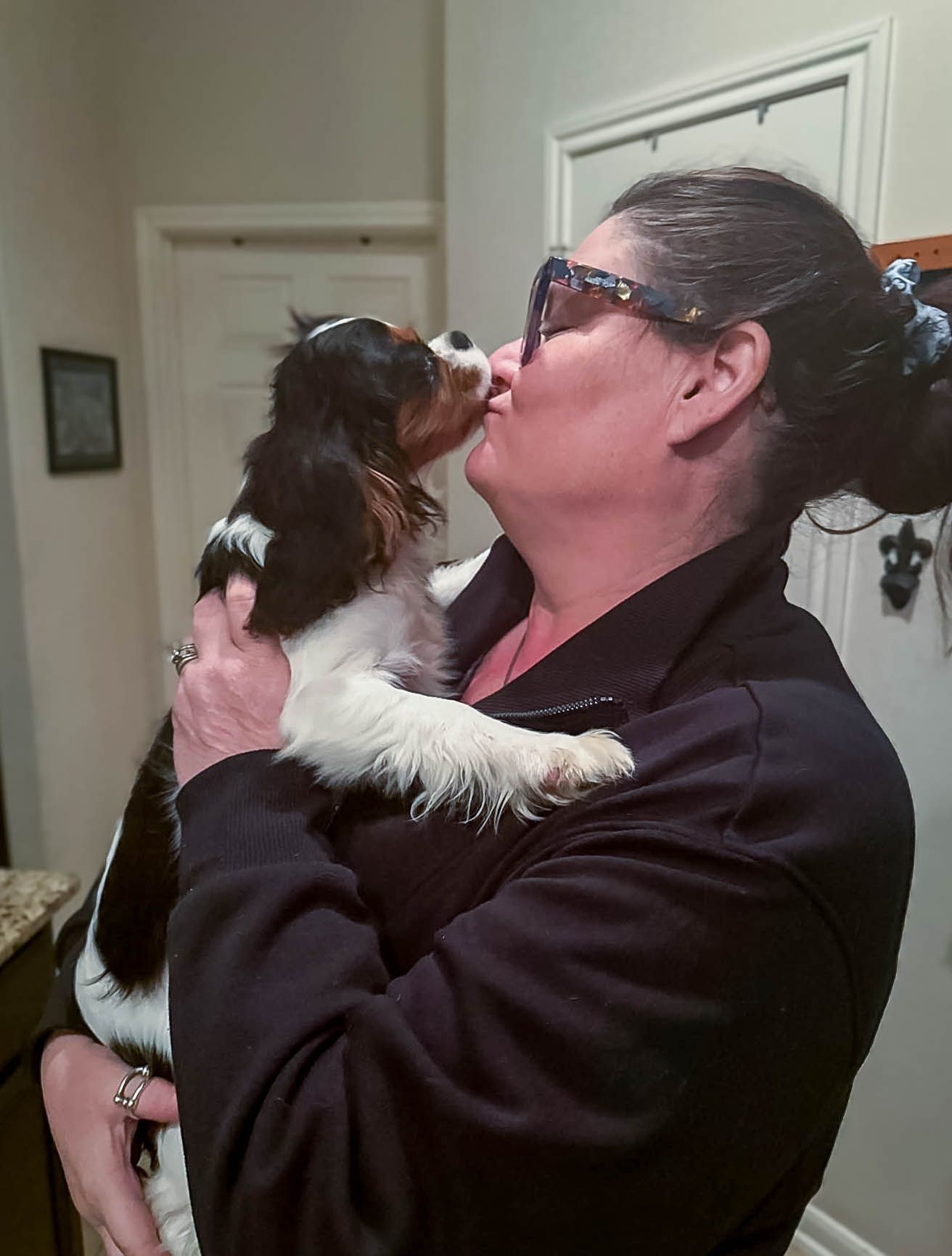 Woman with sunglasses receiving a kiss from a Cavalier King Charles Spaniel puppy she is holding.