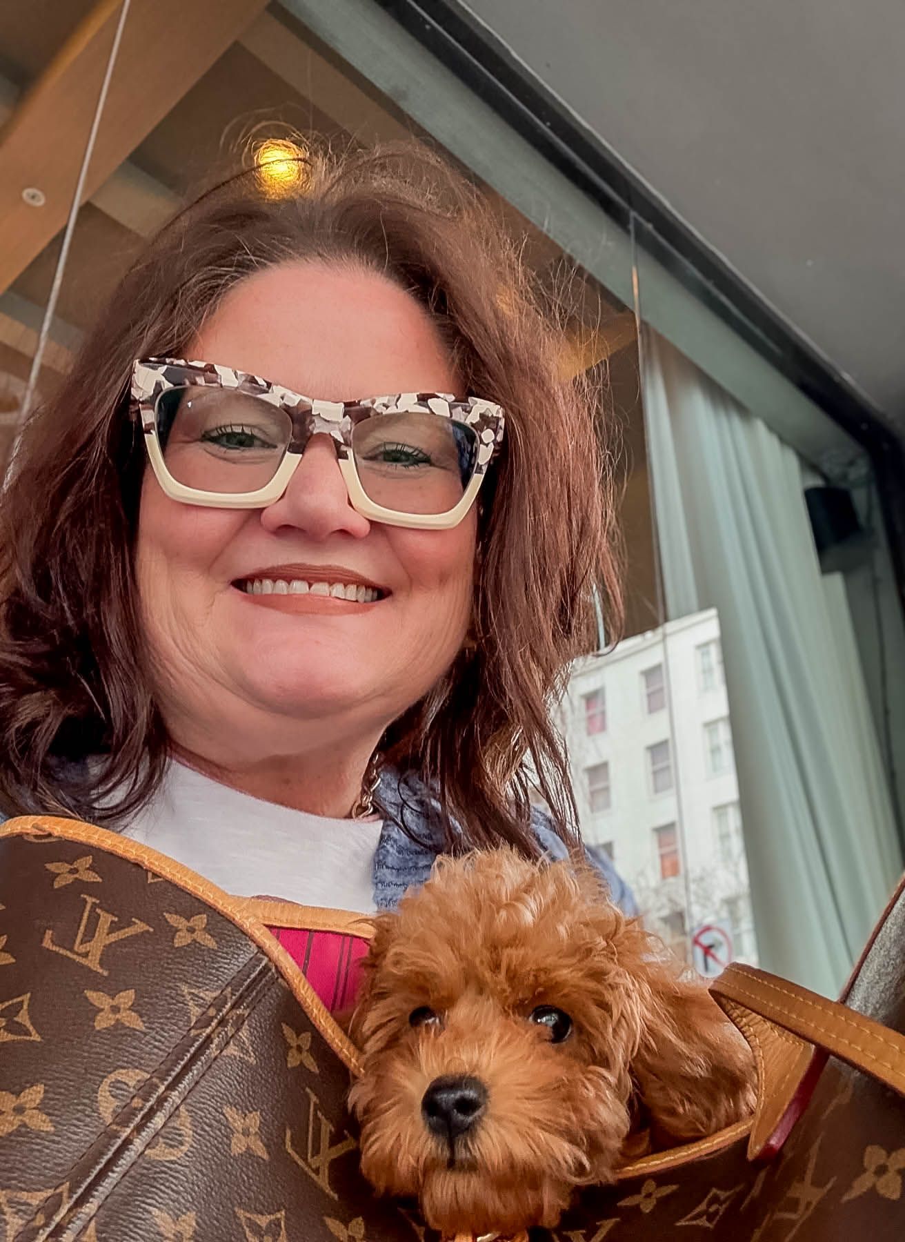 Woman with floral glasses carrying a fluffy red toy poodle in a Louis Vuitton bag during flight nanny layover.