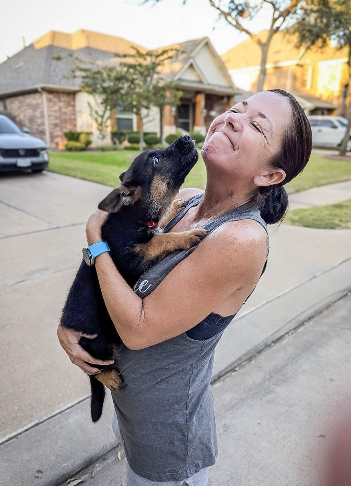 Laughing woman holds a black and tan puppy licking her face on a driveway after pet transport