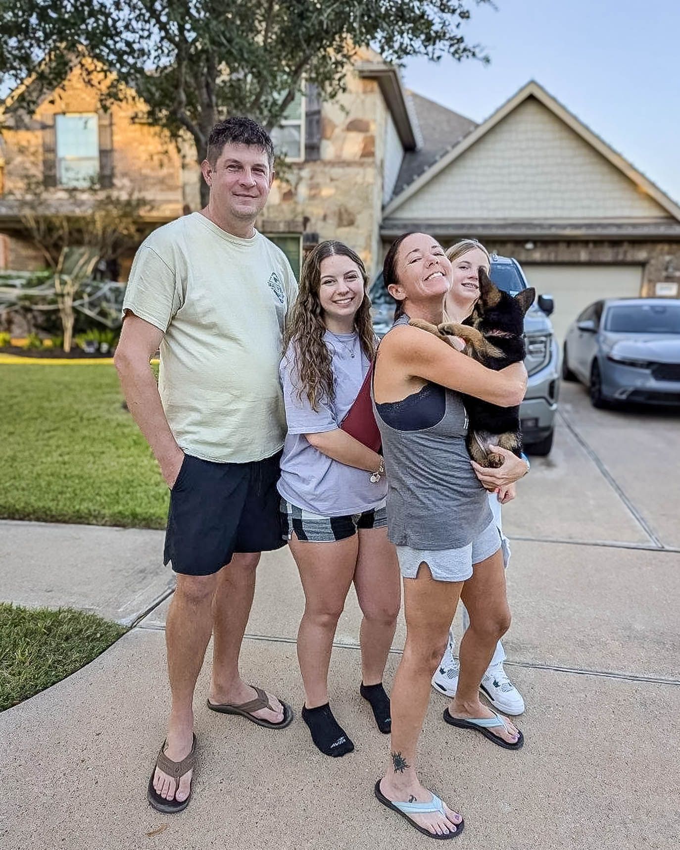 Family of four smiling together in driveway, woman holding small dog, suburban home in background after pet transport.