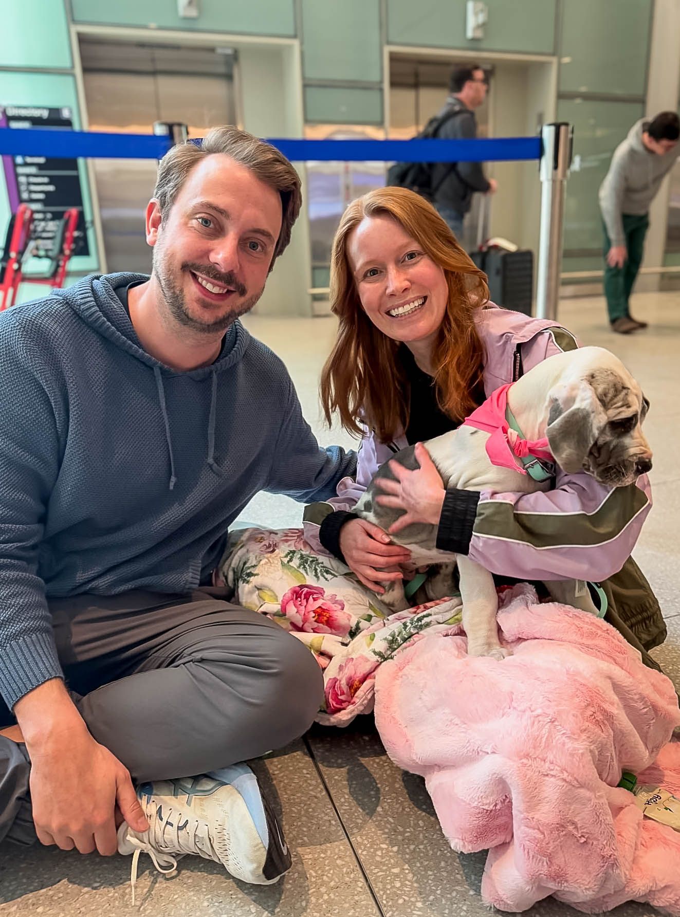 Couple sitting on airport floor smiling with their dog wearing a jacket and pink stuffed animal toy.