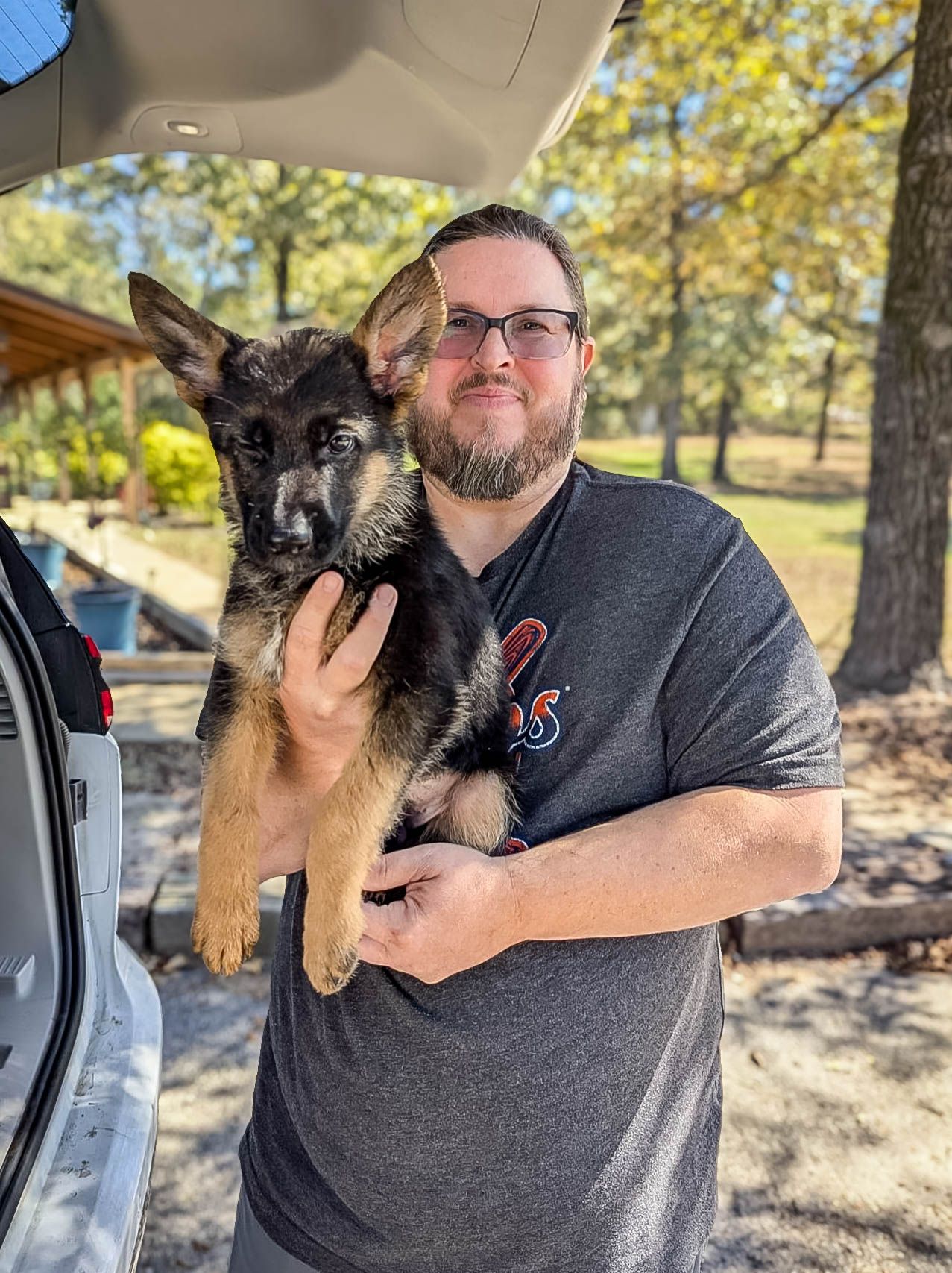 Bearded man in glasses proudly holds a German Shepherd puppy outdoors near trees, after pet transport
